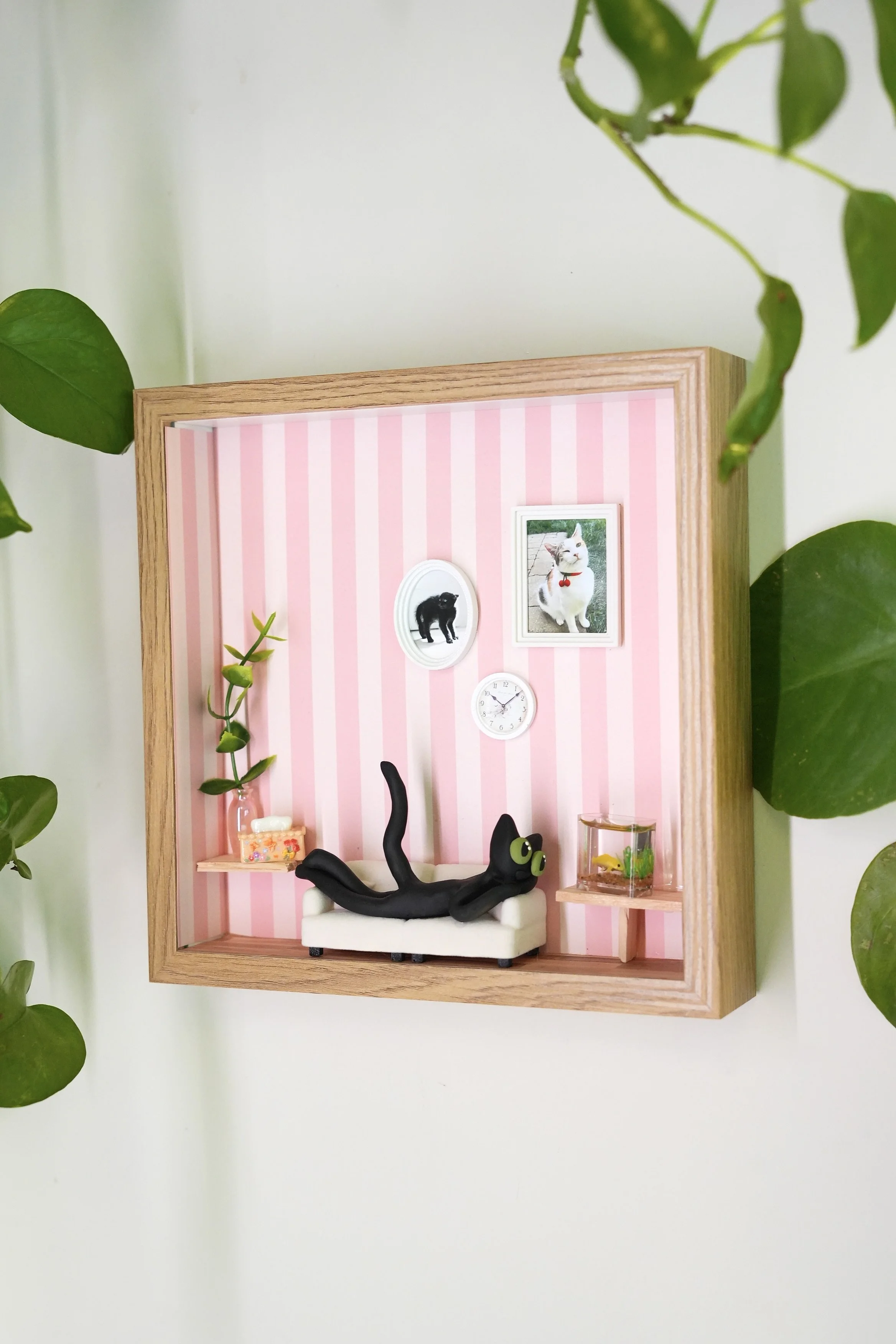 Miniature dollhouse room with pink striped wallpaper featuring framed pictures of a black dog and a white cat, a small white couch with a black cat figure on it, a miniature fish tank, a shelf with a glass of water, and a green plant branch visible a
