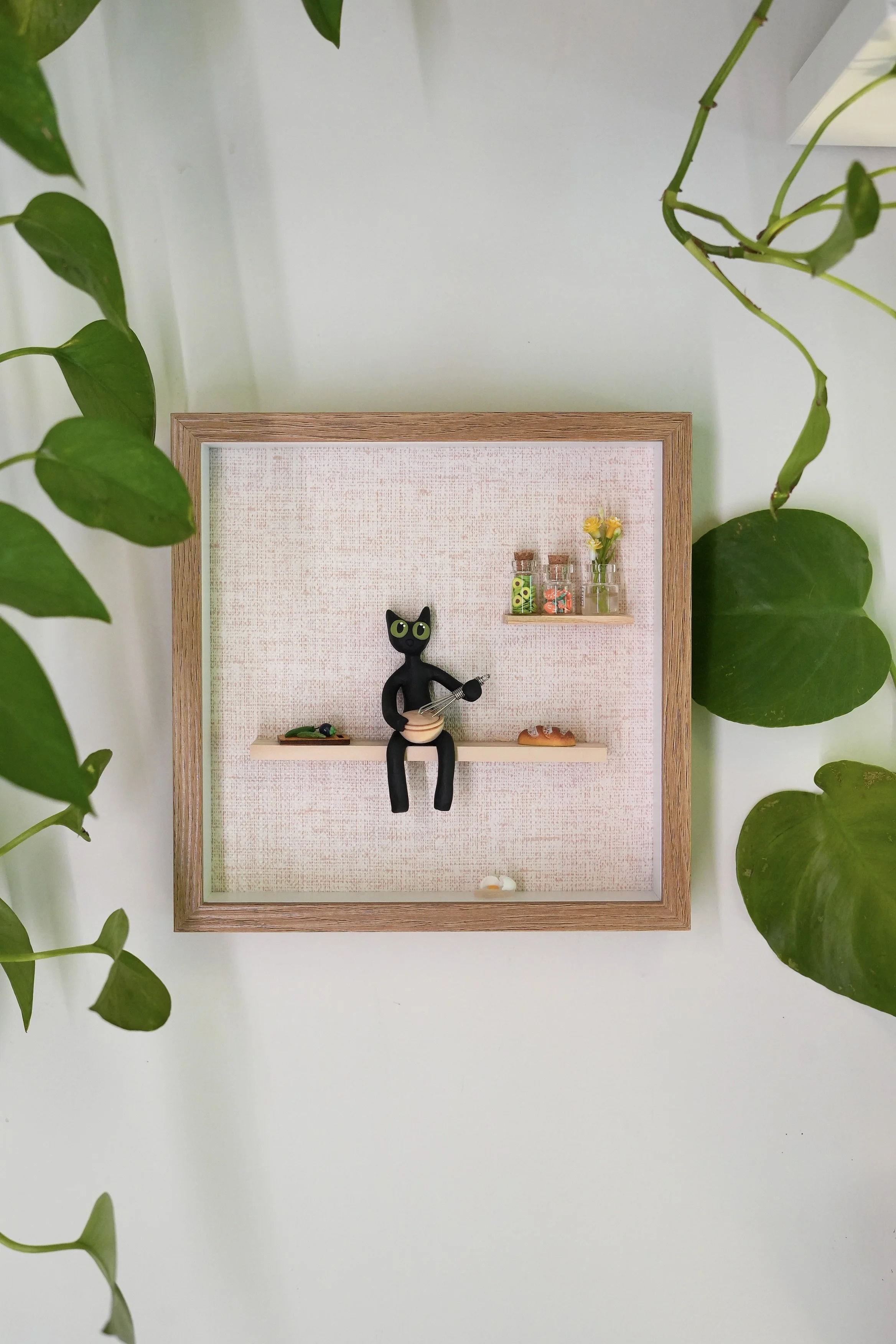Framed display with small decorative items, a black cat figurine holding a whisk, miniature plants, and a small piece of bread, surrounded by green leafy plants on a white background.