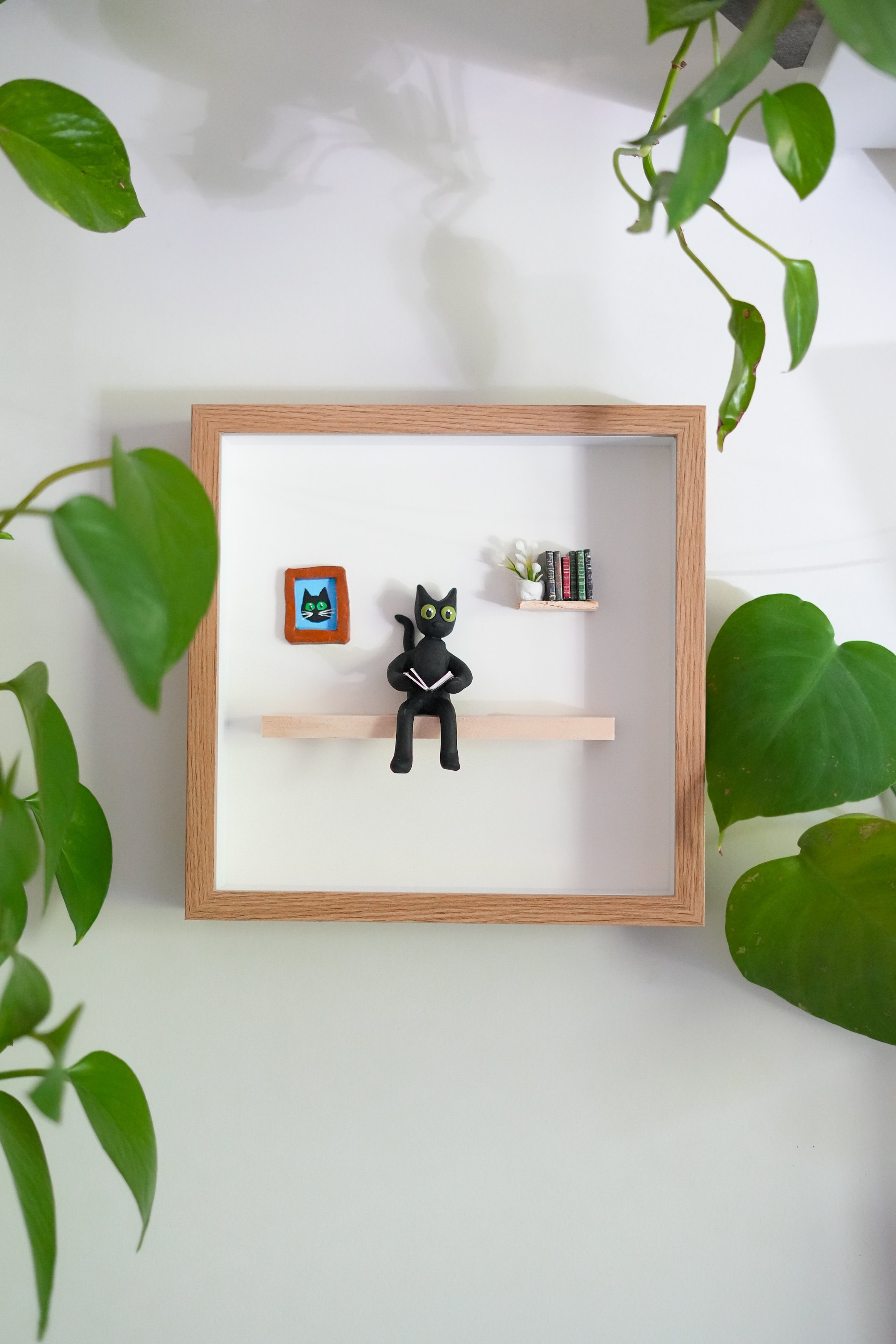Decorative wall frame with small black cat figurine, miniature bookshelf with books, and framed picture, surrounded by green plant leaves.