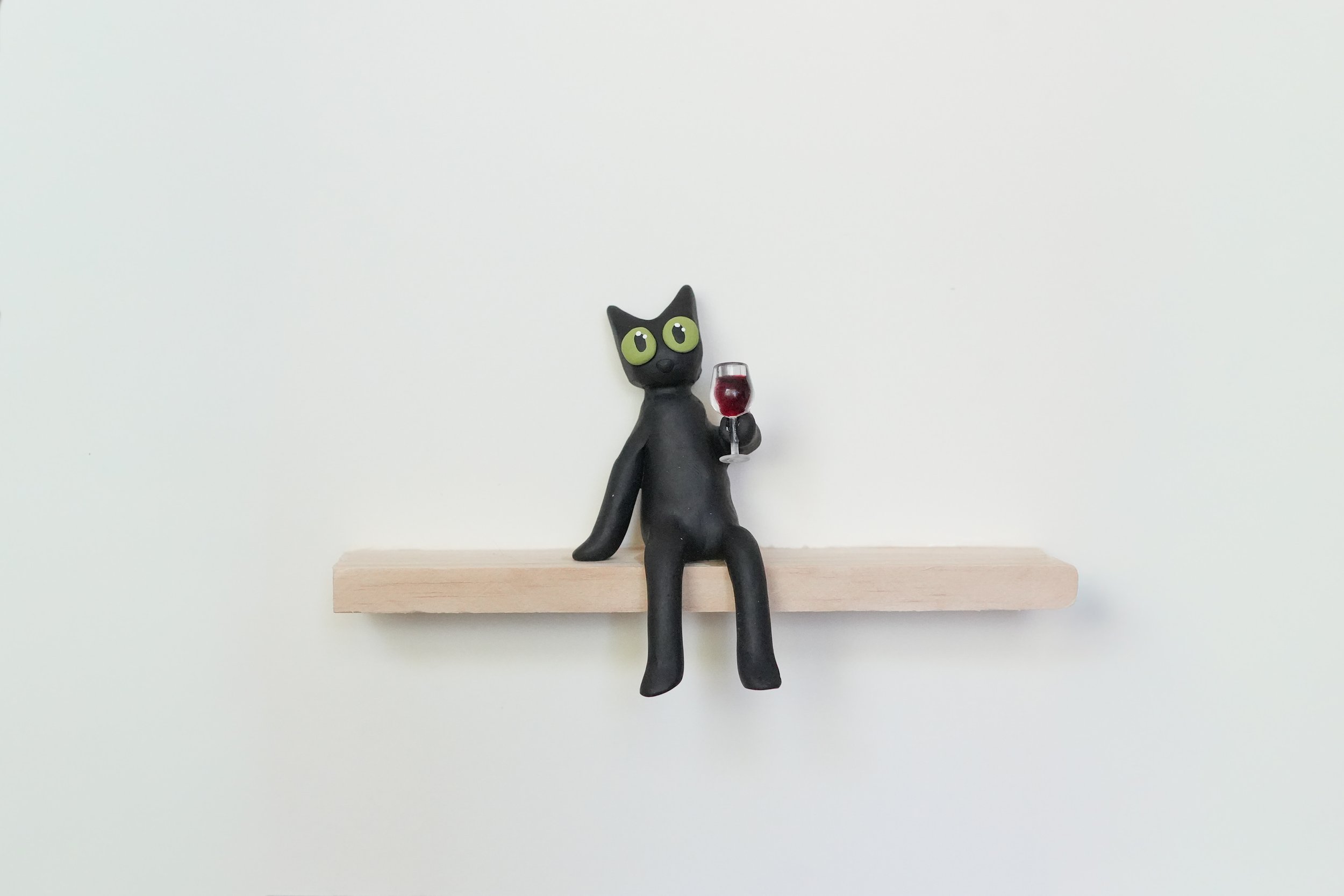 Black cat figurine with large green eyes sitting on a wooden shelf, holding a glass of red wine, against a plain white background.