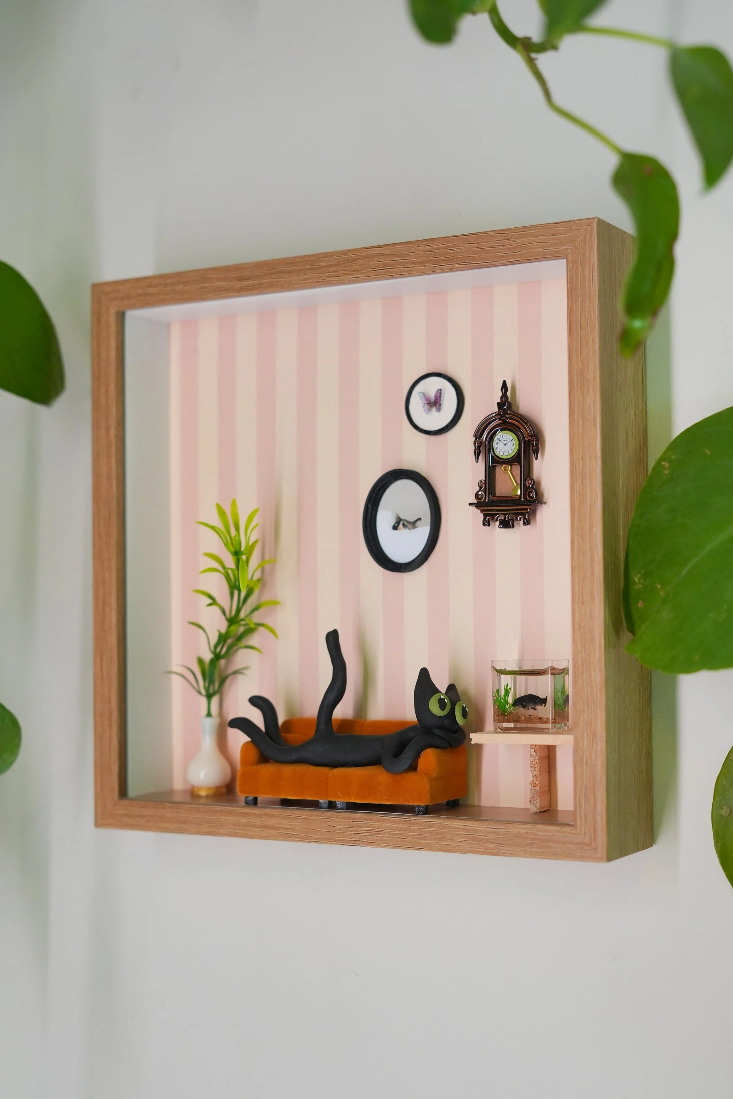 Miniature diorama of cozy living room scene with black cat figurine sleeping on orange sofa, green plants, framed butterfly and bird pictures, vintage clock, and small fish tank, surrounded by green houseplants.