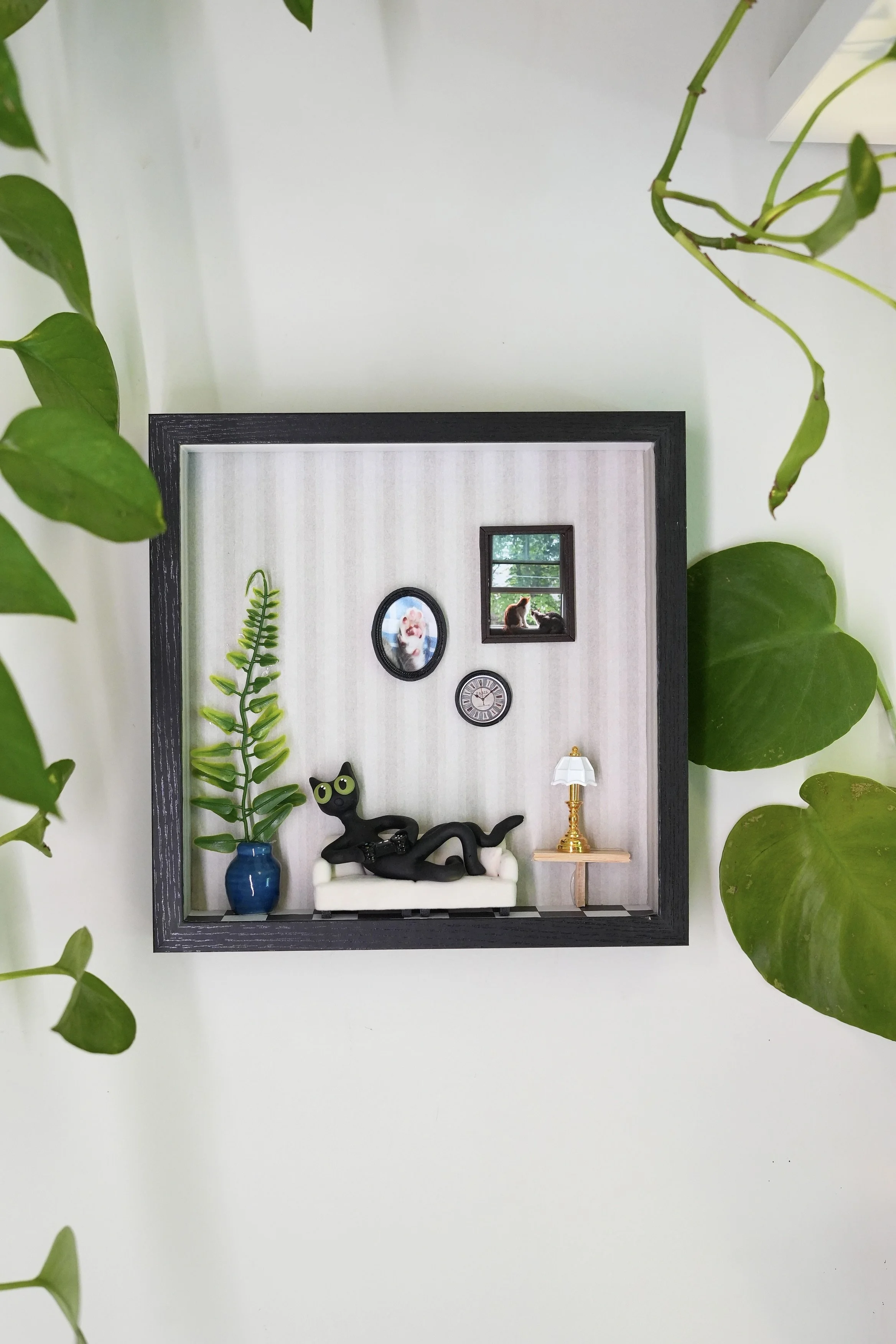 Decorative wall display with framed pictures, a black cat figurine, a small lamp, a picture of a dog and cat on a window, a green plant, and surrounding green leaves.
