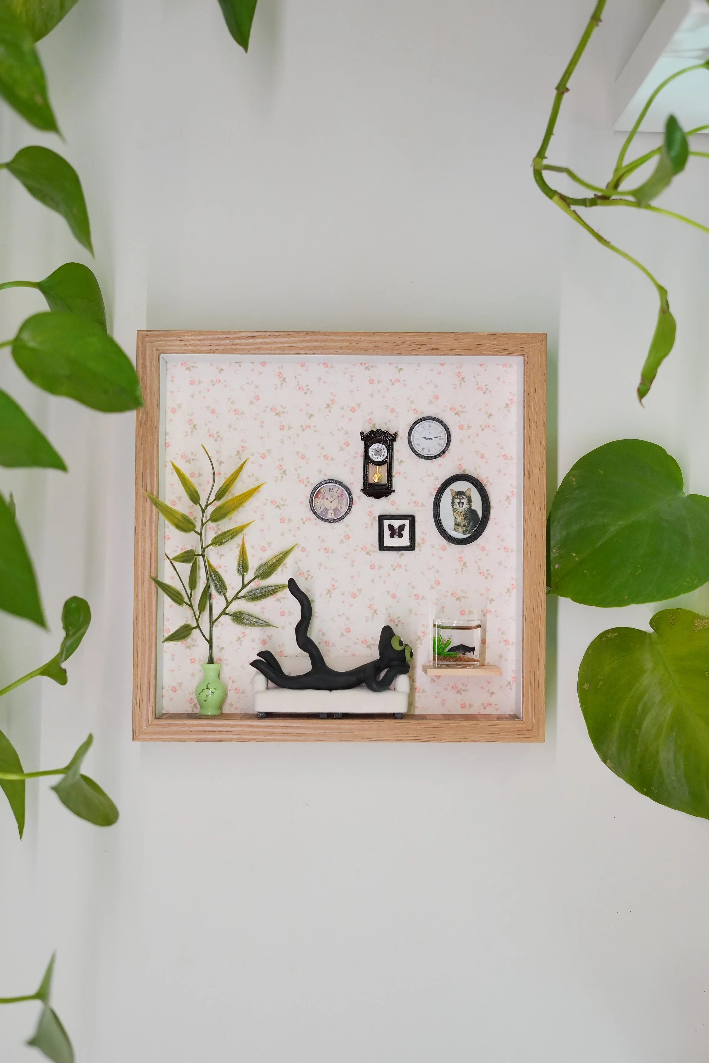 Decorative picture frame with small garden-themed objects, greenery, and clock, surrounded by green plant leaves.