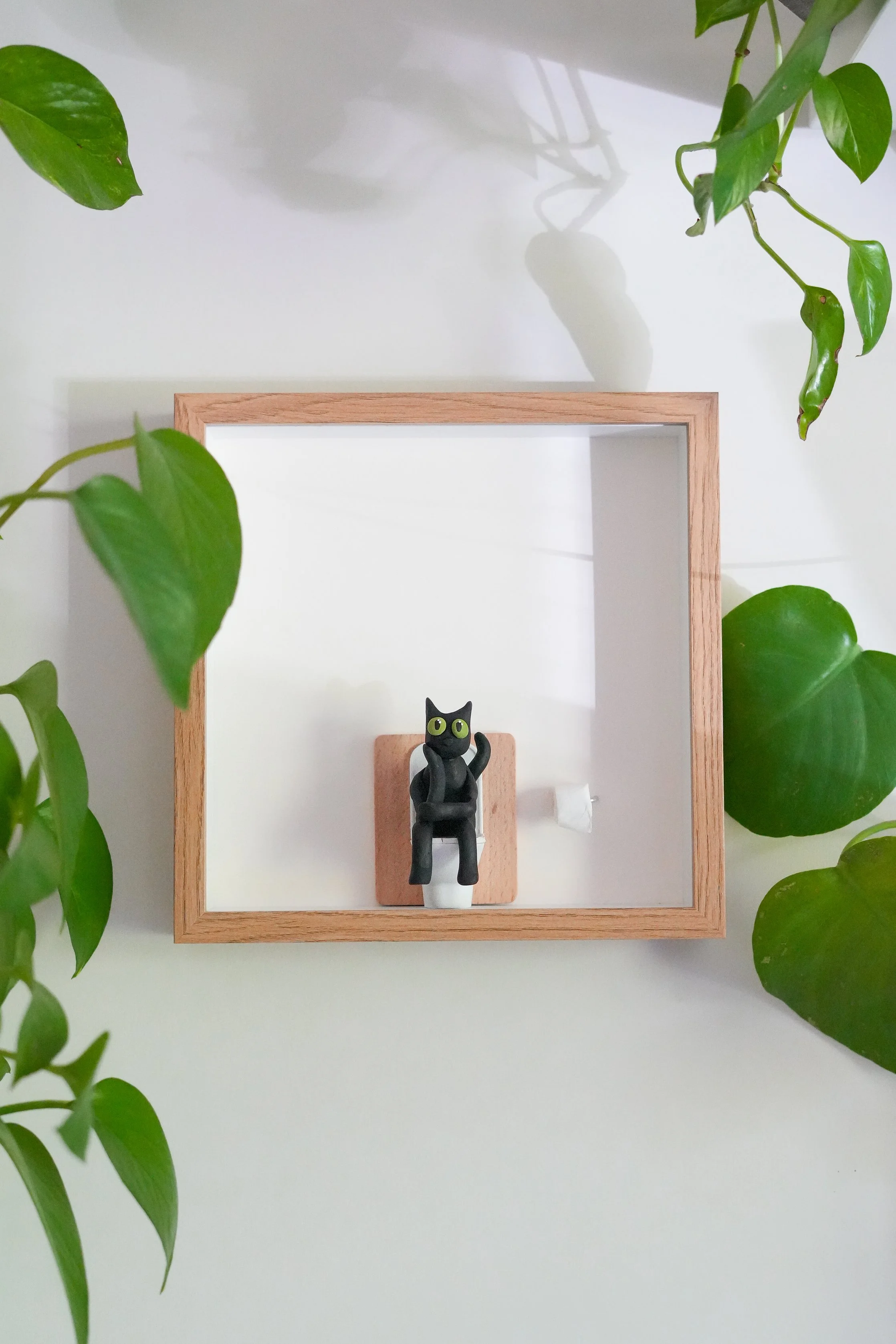 A black cat figurine with green eyes sitting inside a wooden frame mirror, surrounded by green leafy plants on a white surface.