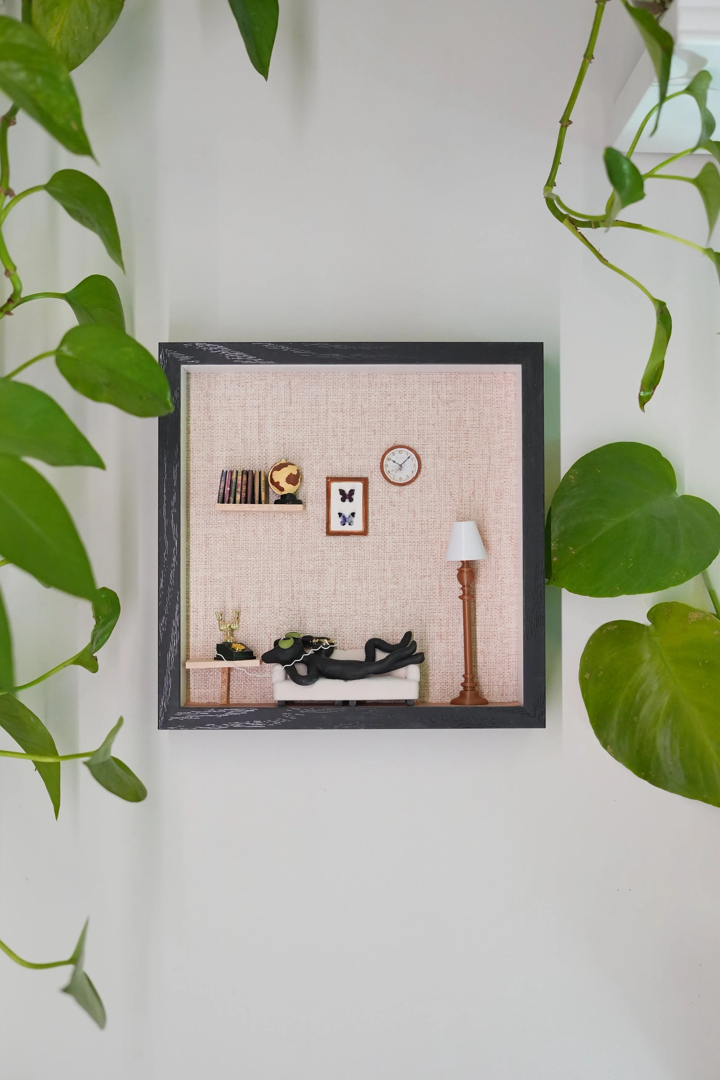 Decorative wall shelf with a globe, books, a butterfly picture, a clock, and some decorative items including a figure on a small sofa, a lamp, and plants framing the scene.