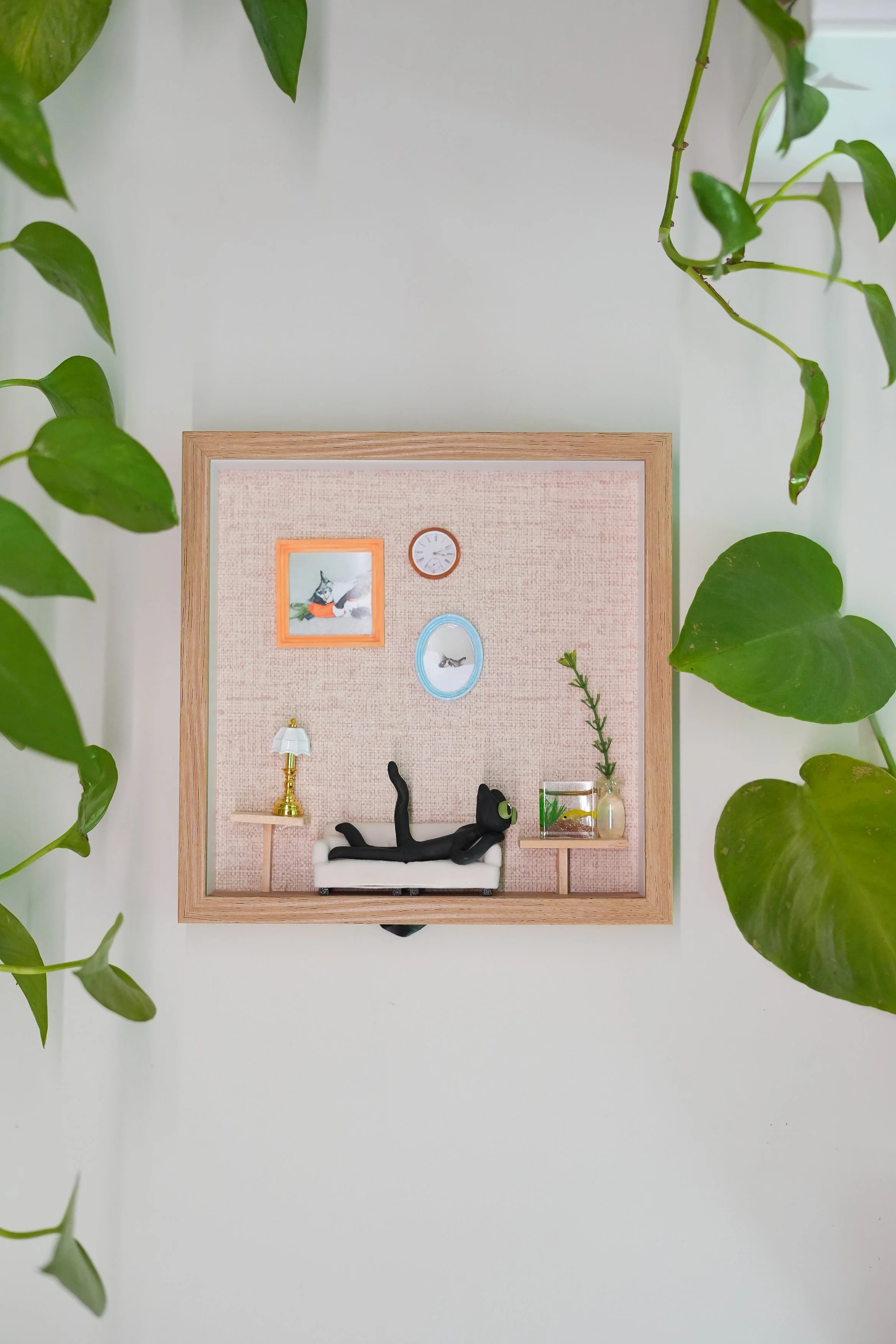 Miniature room scene with a small black cat lying on a couch, a side table with a lamp, and framed pictures on the wall, surrounded by leafy green plants.