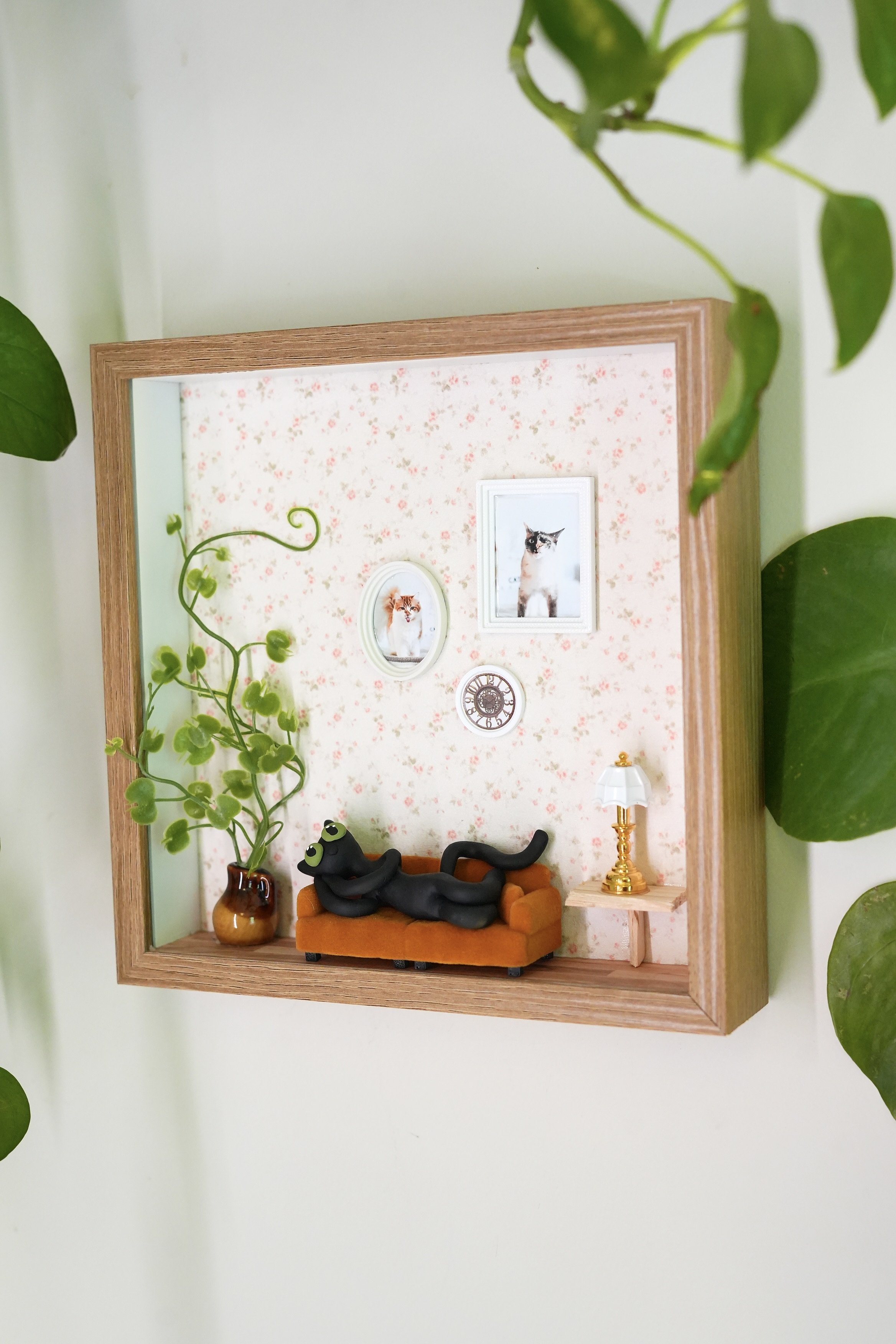 Miniature wall display with framed kitten photographs, a small table lamp, a potted plant, and a black cat sculpture lounging on an orange sofa.