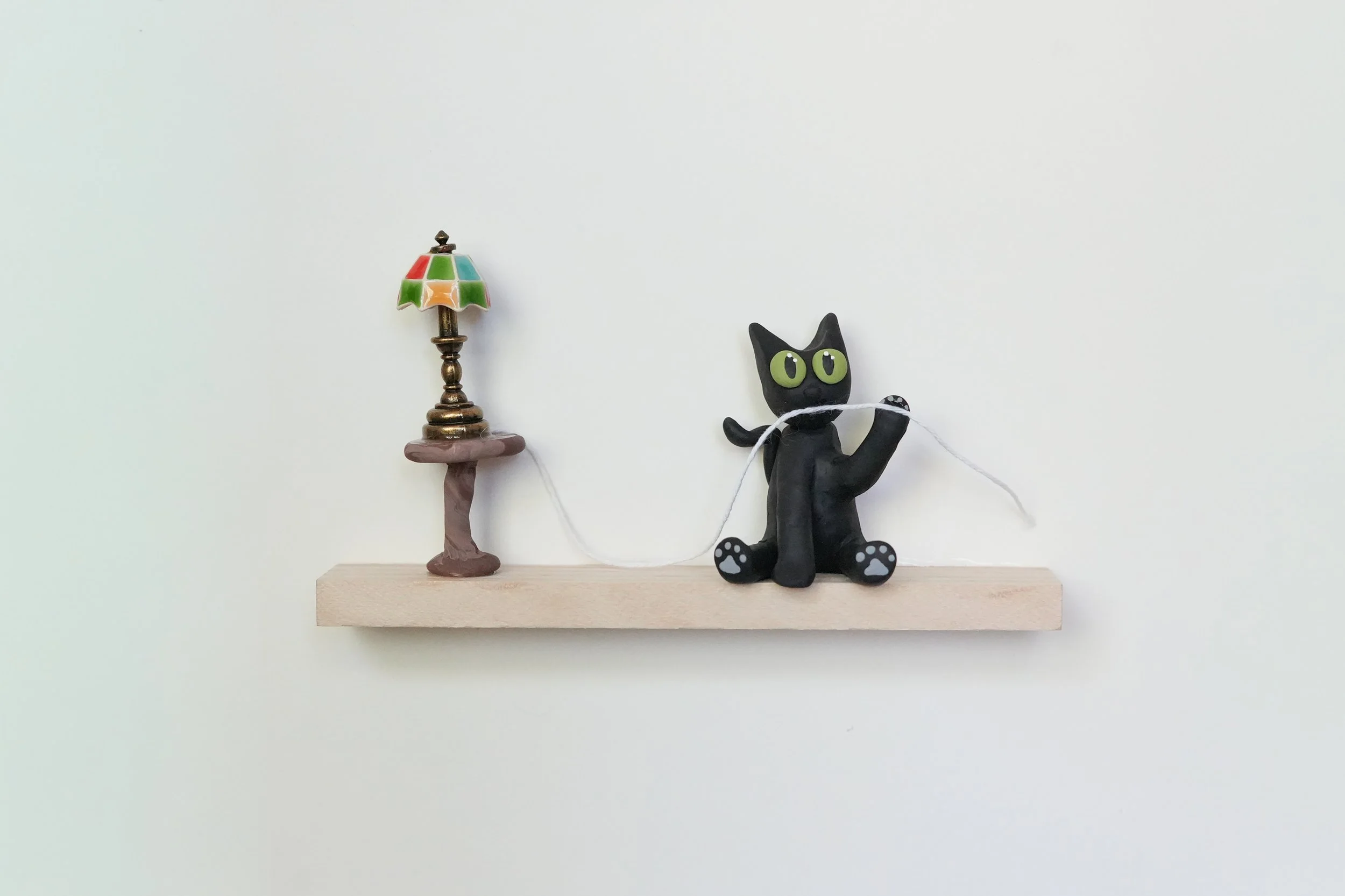 A small wooden shelf mounted on a plain white wall, holding a miniature lamp with a stained glass shade and a black cat figurine made of clay or similar material with green eyes, sitting with one paw raised.