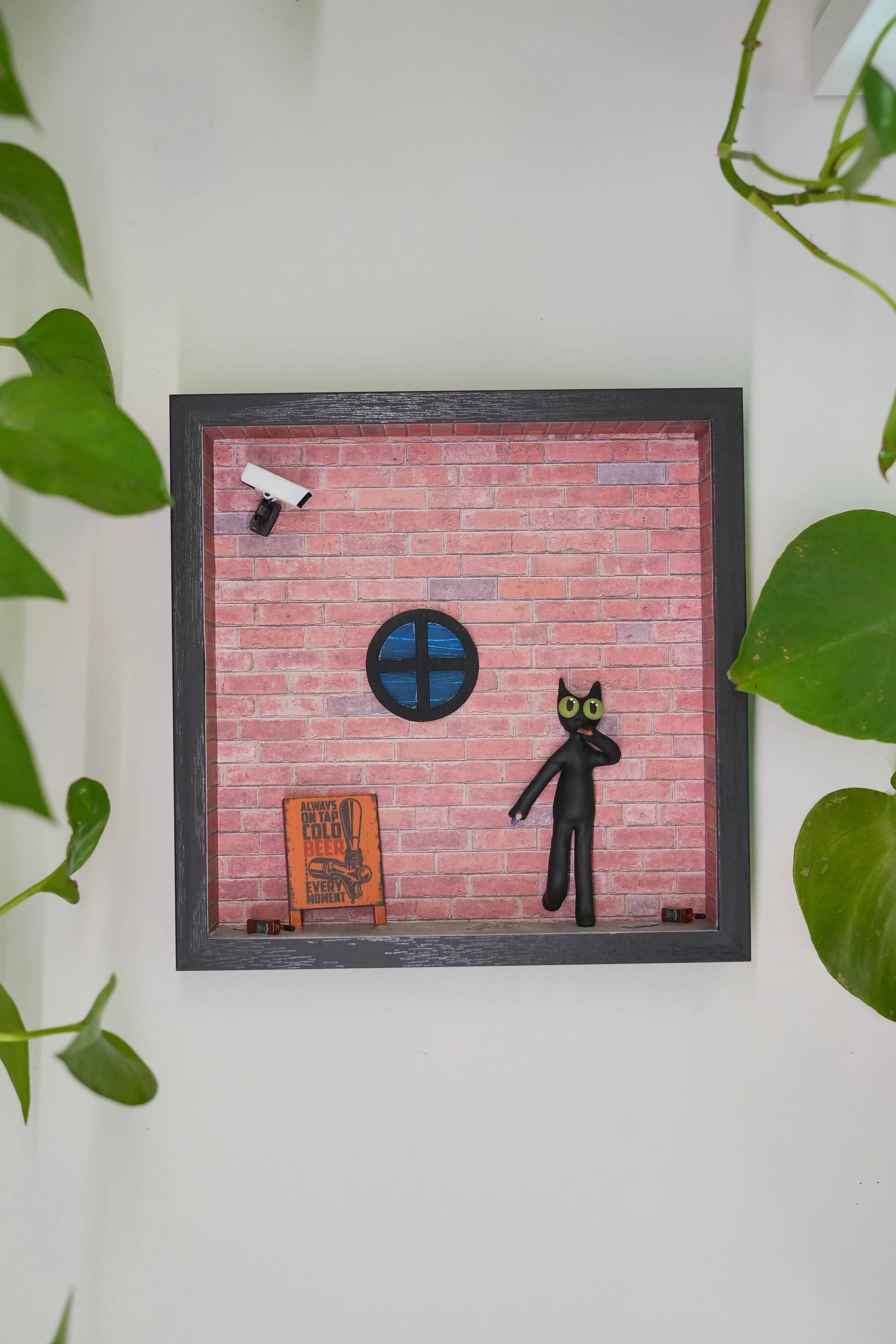 A shadow box with a red brick wall background, a small black cat figurine with green glasses holding a purple pencil, a miniature orange sign reading 'Always on tap, cold beer, moment!', a small orange and black sign, and two small clip lights at the