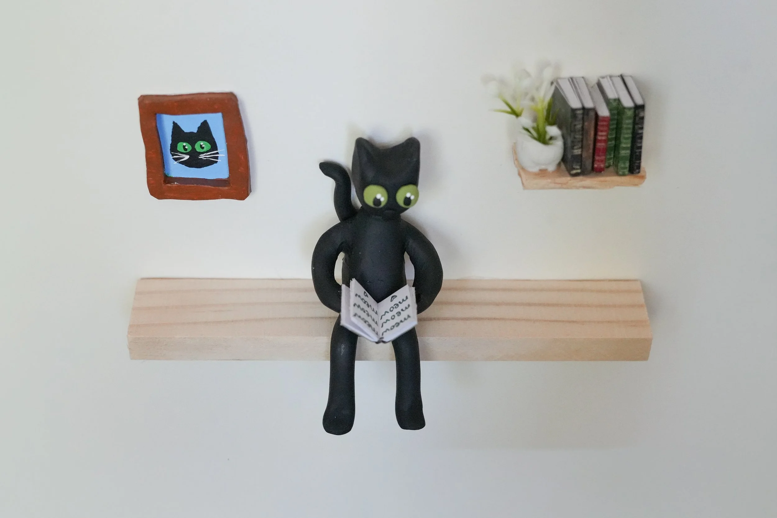 A black cat figurine with large green eyes sitting on a wooden shelf, holding an open book, with a small framed picture of a black cat and a bookshelf with books and a plant on the wall behind.