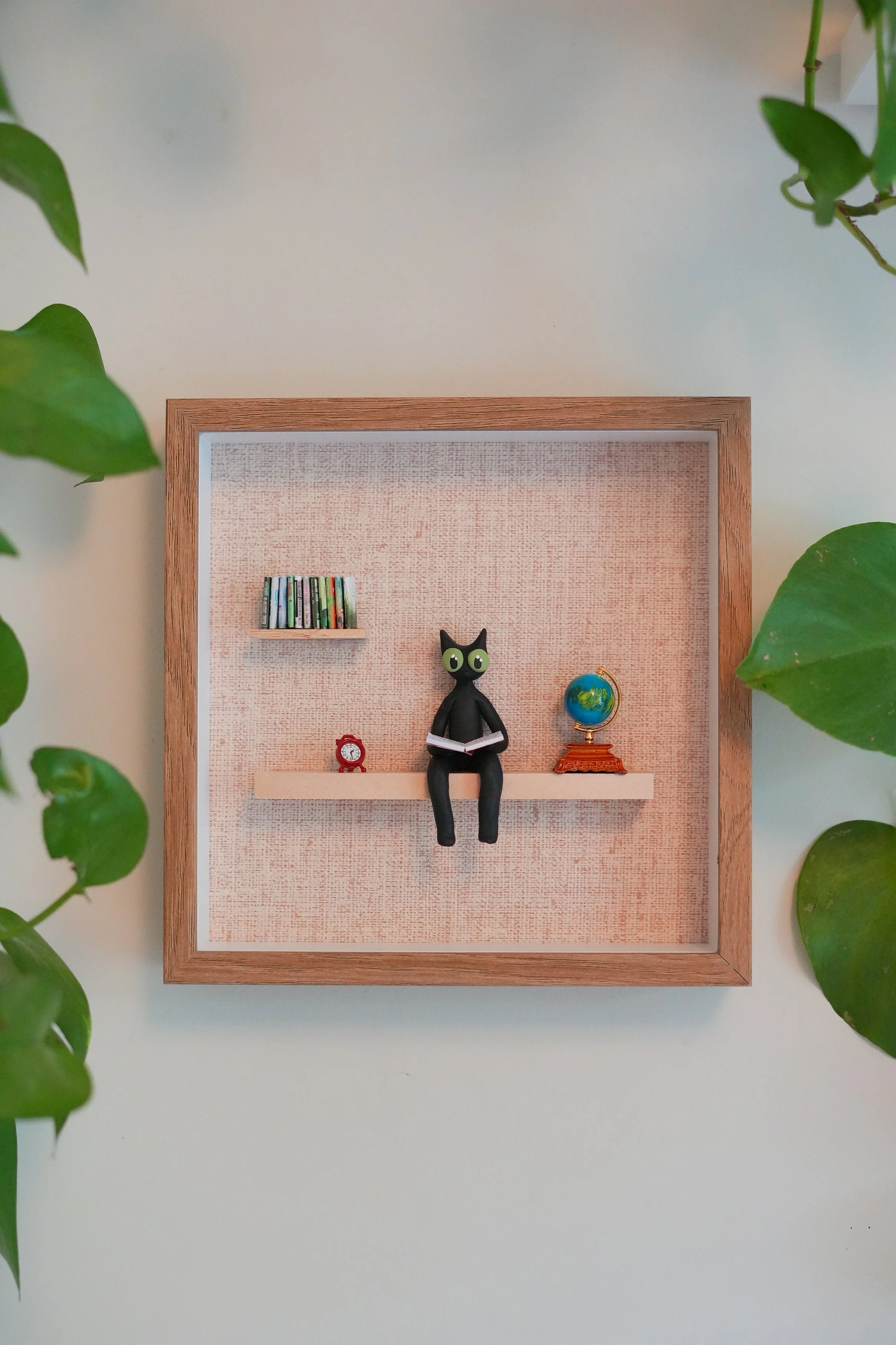 Decorative framed wall with small bookshelf, black cat figurine reading a book, globe, and red alarm clock, surrounded by green leaves.