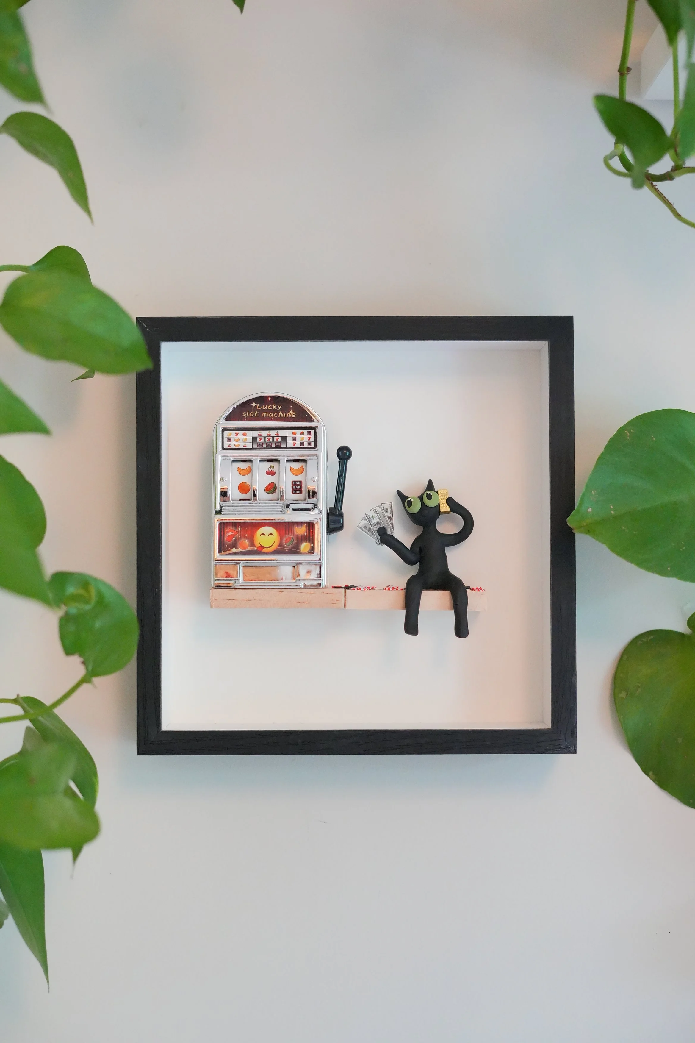 Decorative wall display with a miniature slot machine and a black cat figurine holding money, surrounded by green leafy plants on a white wall