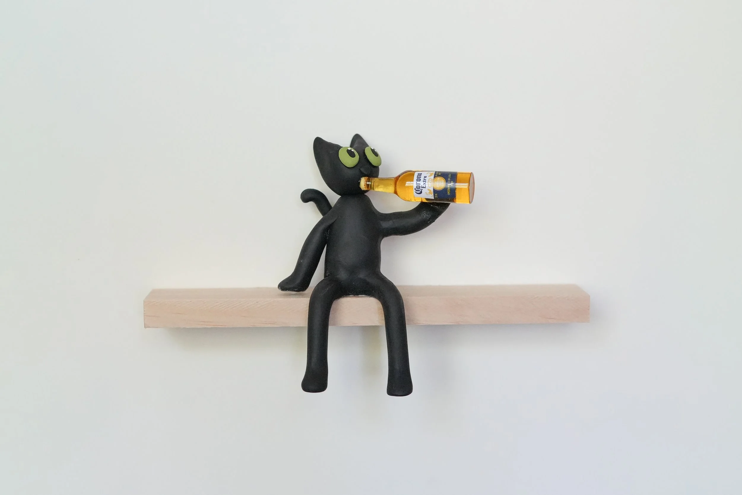A black cat figurine with green eyes sitting on a wooden shelf, holding a small Corona beer bottle in its paw.