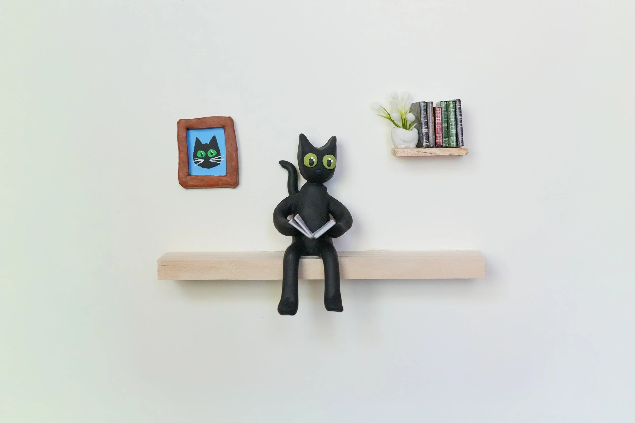Miniature black cat figurine reading a book, with a framed cat picture on the wall, a small bookshelf with books and a white vase with flowers on a plain white wall.