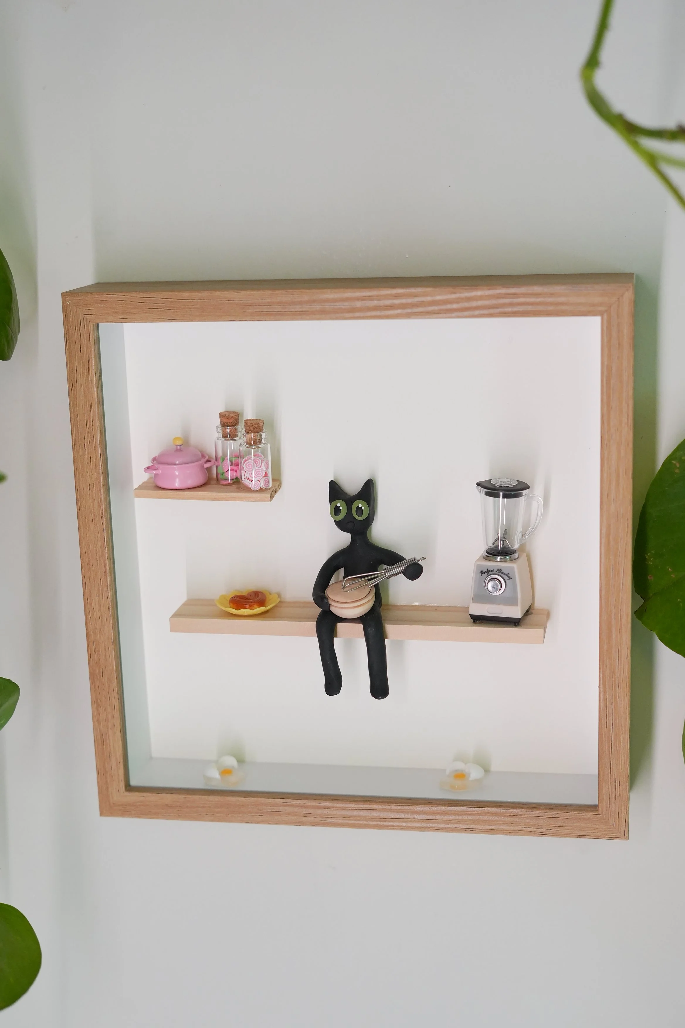 Decorative wall shelf with kitchen-themed items, including a pink teapot, small jars with cork lids, a black cat figurine holding utensils, a yellow plate with what appears to be soap or small objects, and a blender.