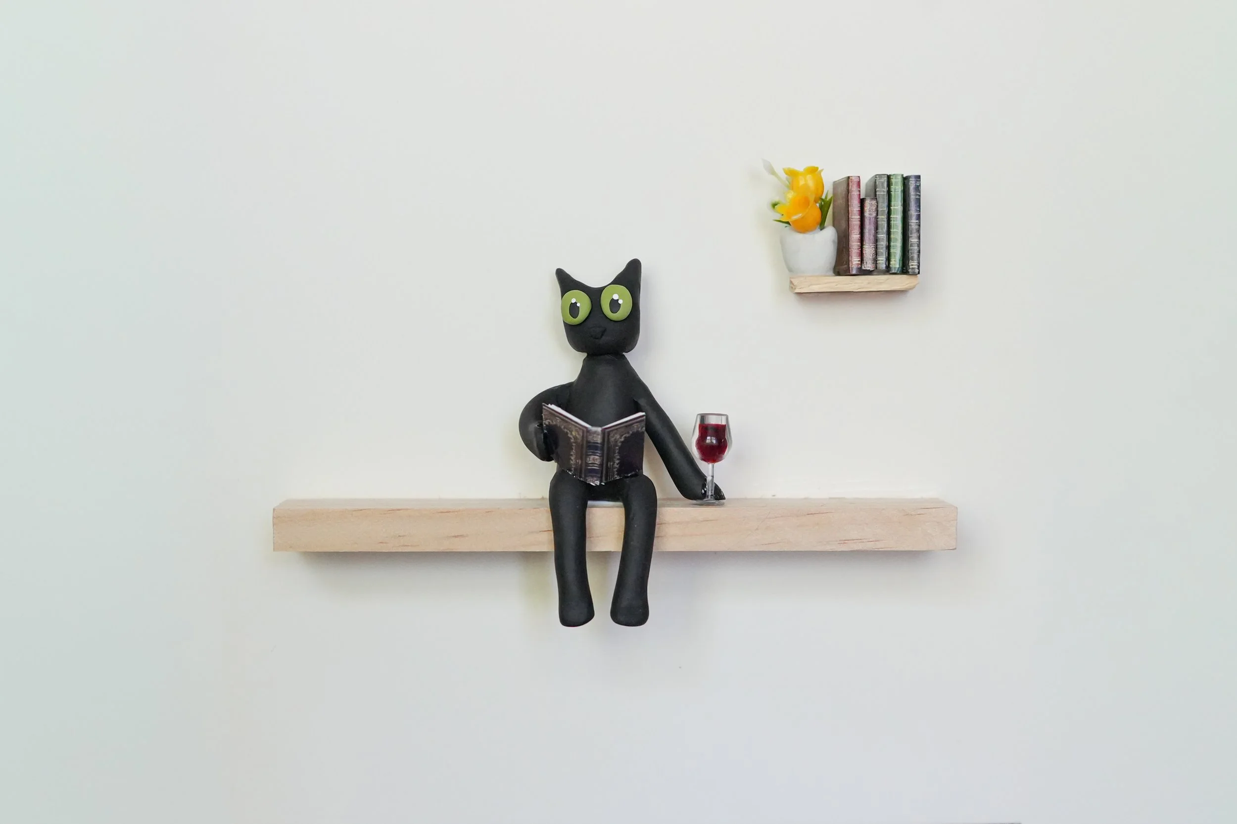 A decorative black cat figurine with large green eyes, sitting on a wooden shelf, holding a book and a glass of red wine. There is a second small shelf on the wall with a white pot containing yellow flowers and a row of colorful books.