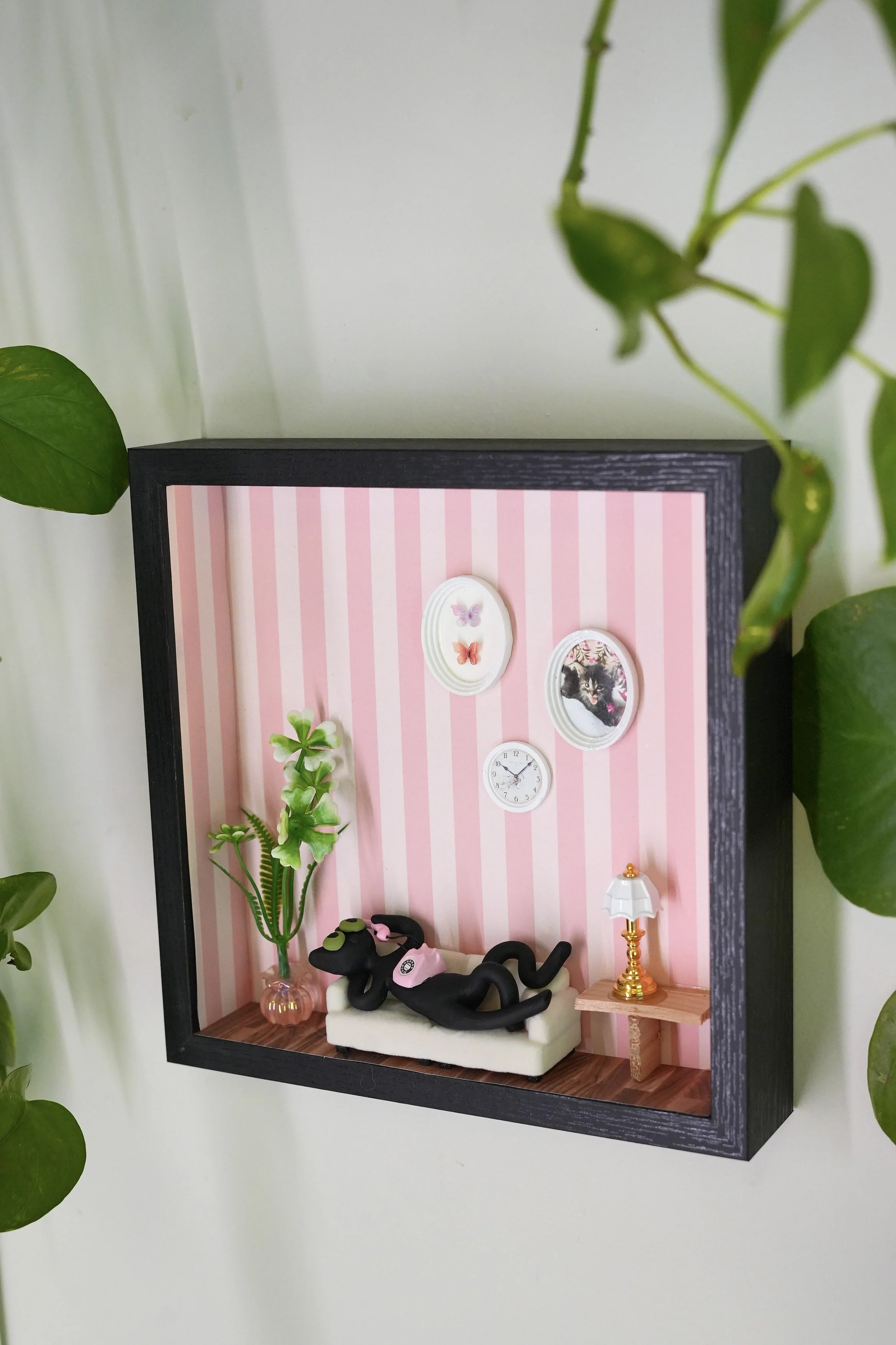 Miniature dollhouse room with pink striped wallpaper, potted plant, black cat figurine, small sofa, and a side table with a gold lamp, surrounded by green houseplants.