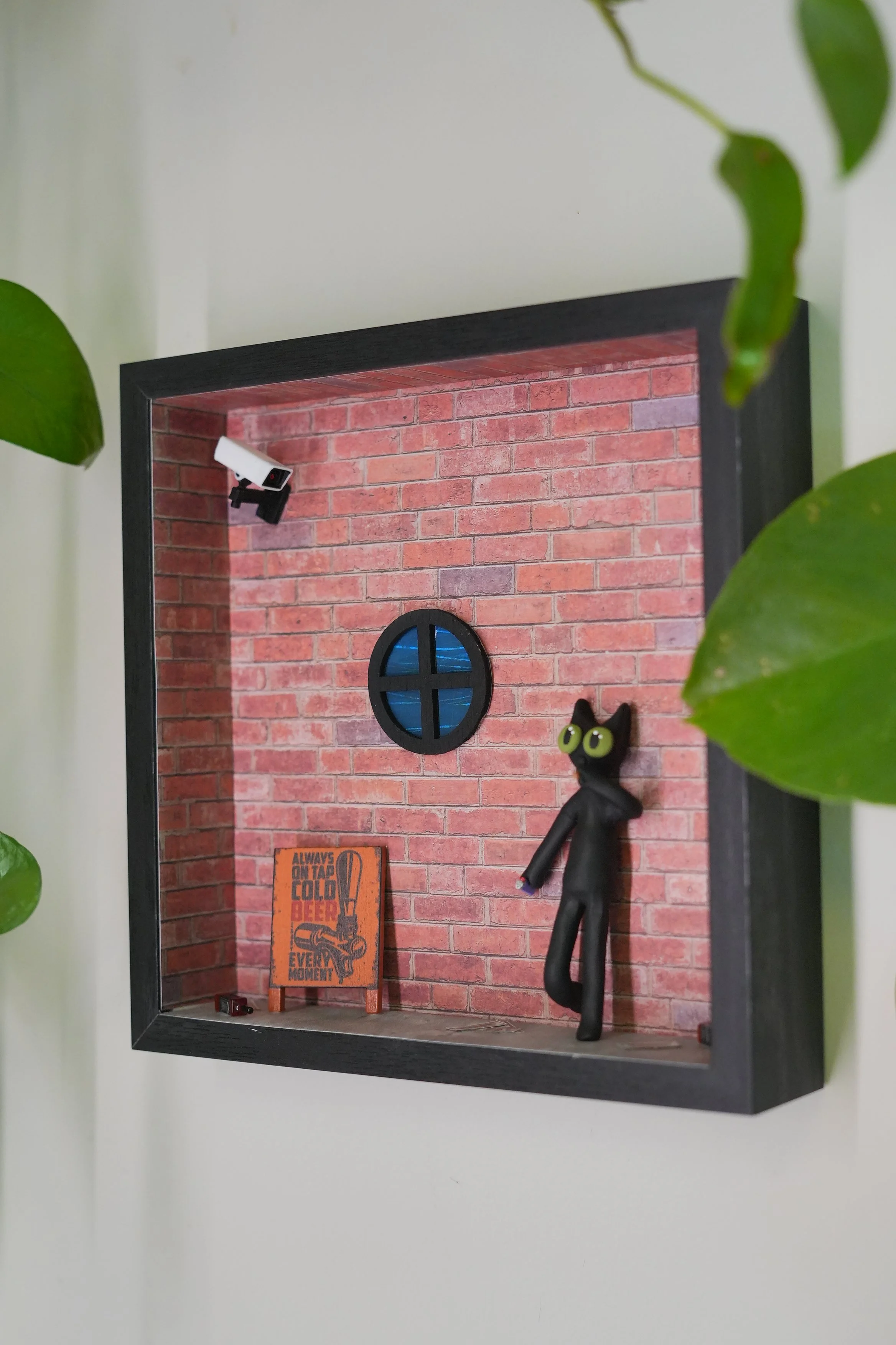 Miniature room with red brick wall background, black window, black cat figure with green eyes, security camera in the top left corner, and a sign on the floor that reads 'Always on tap cold beer every moment', with green leaves partially visible at t