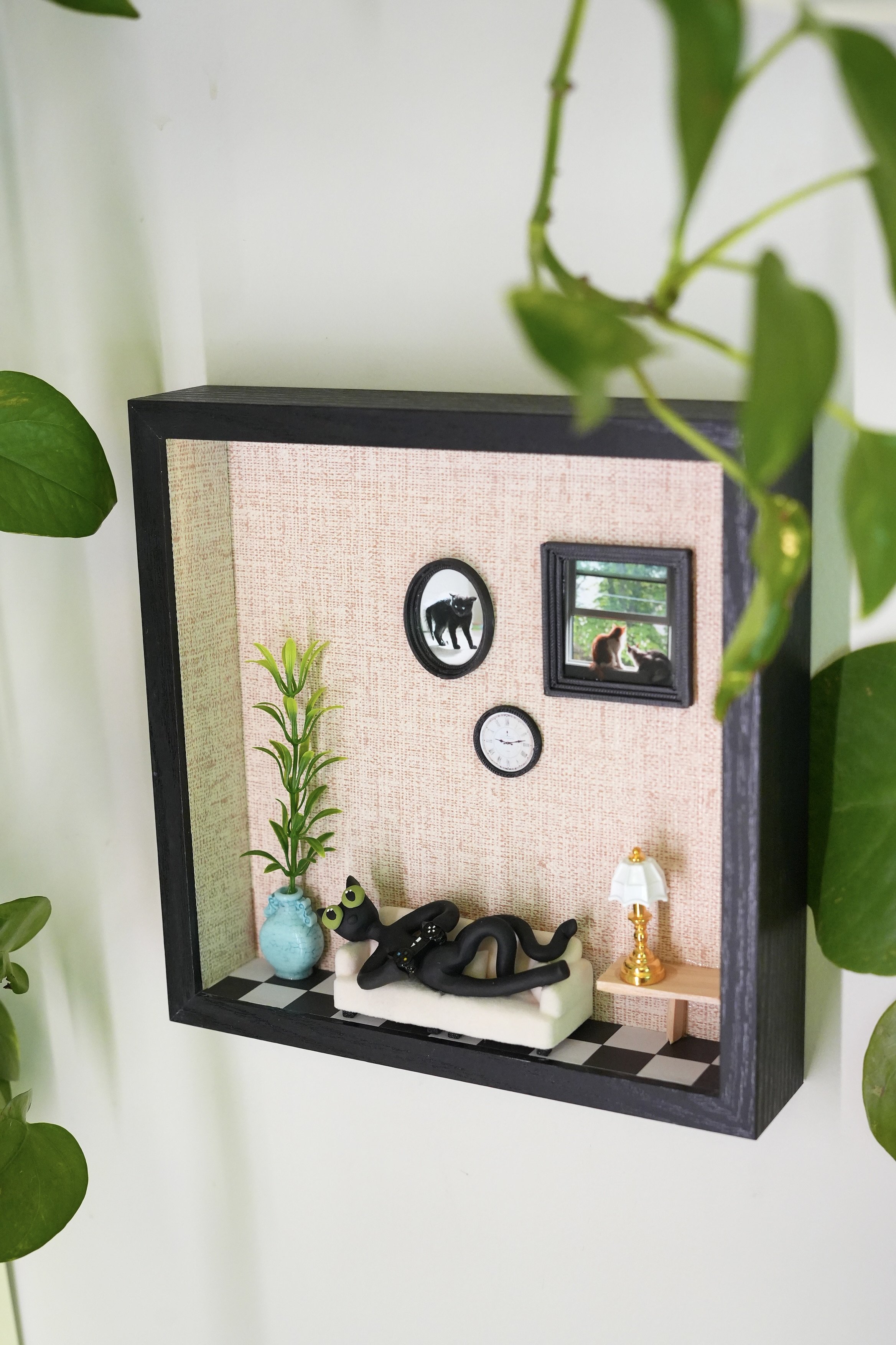 A miniature living room scene with pink patterned wallpaper, a black framed oval mirror reflecting a black cat figurine, and a window showing an outdoor view. Decor includes a tall green plant in a blue vase, a white sofa with a black cat figurine, a