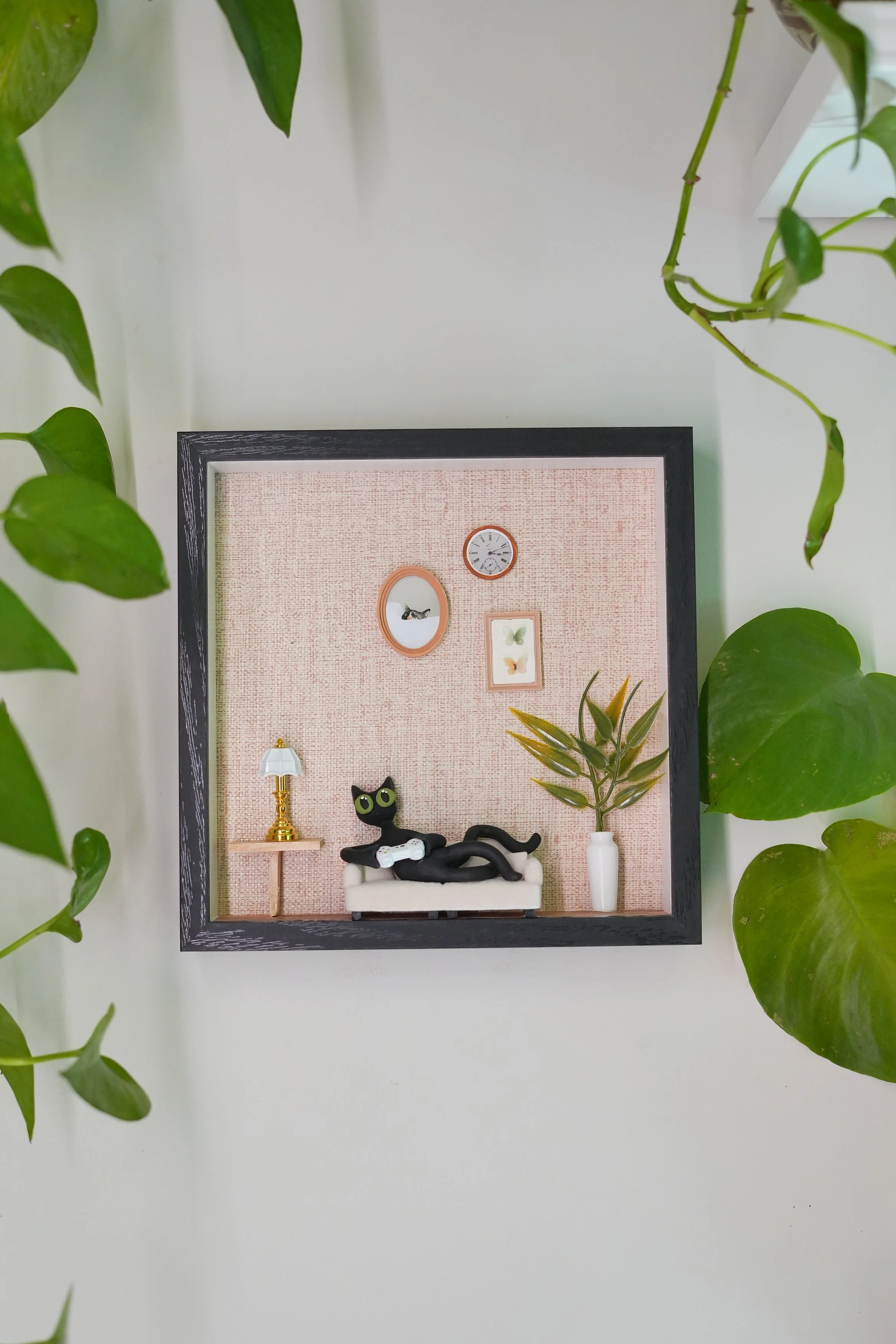 Miniature framed diorama with a black cat figurine, a gold lamp, a vase with a plant, a clock, and pictures of butterflies on a pink textured background, surrounded by green plant leaves.