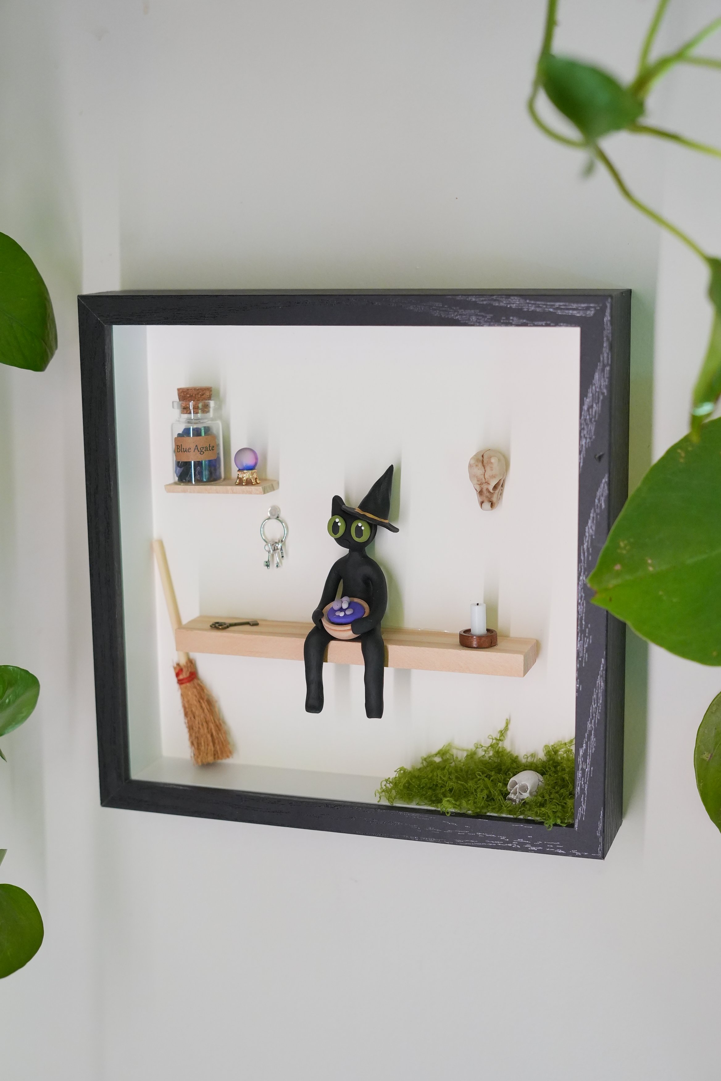 Decorative shelf with a small broom, a glass bottle labeled 'Blue Agate', a small globe, a witch's hat with green eyes, a skull, and some green moss, surrounded by green leaves.