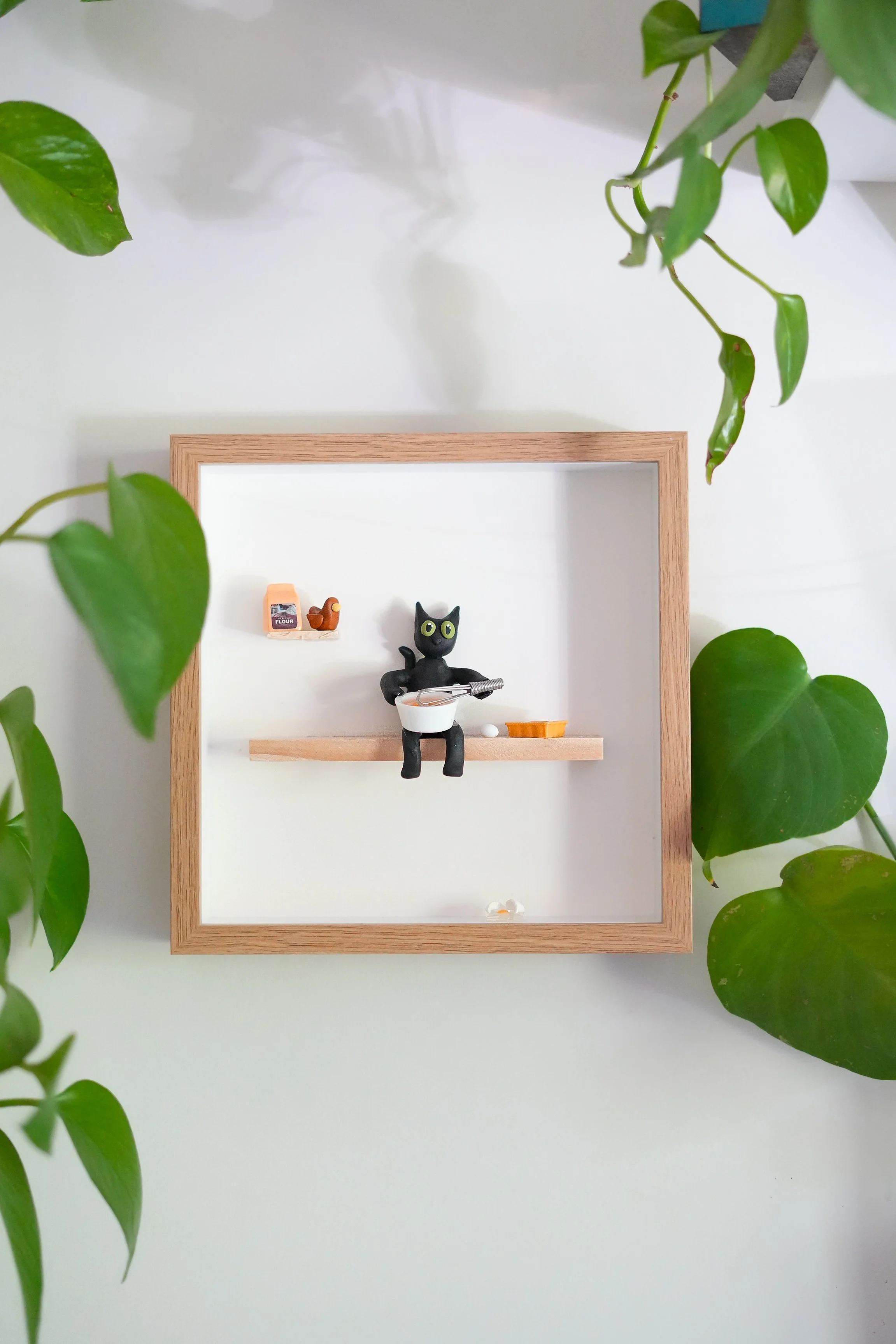 Decorative shadow box with a small black cat figurine holding a white bowl, a miniature orange tray, a tiny bookshelf with a book, and small food items inside. Green foliage partially surrounds the shadow box.