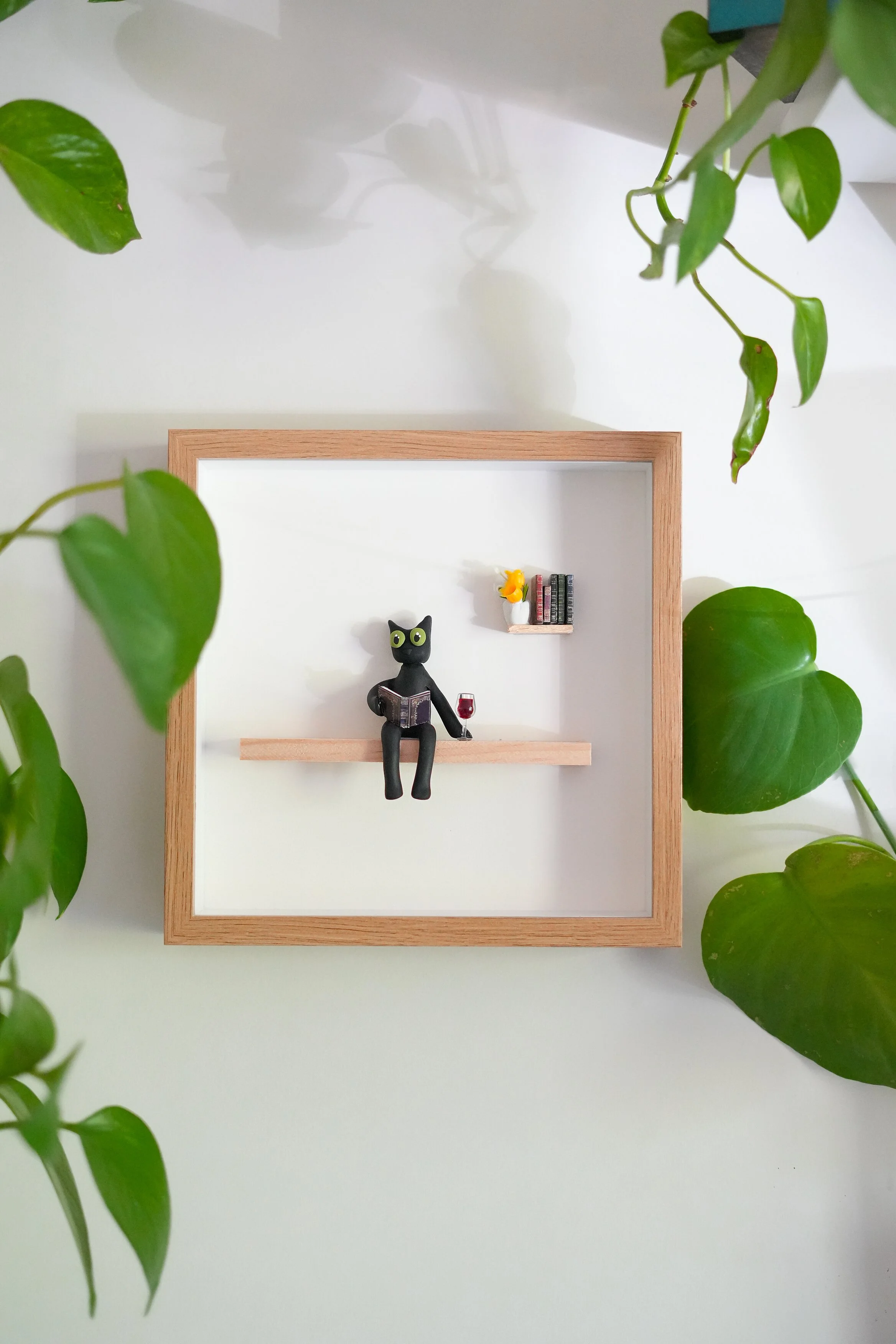 Decorative shadow box with a small black cat figurine reading a book, a miniature wine glass, a small shelf with books, and a tiny vase with yellow flowers, surrounded by green houseplants.