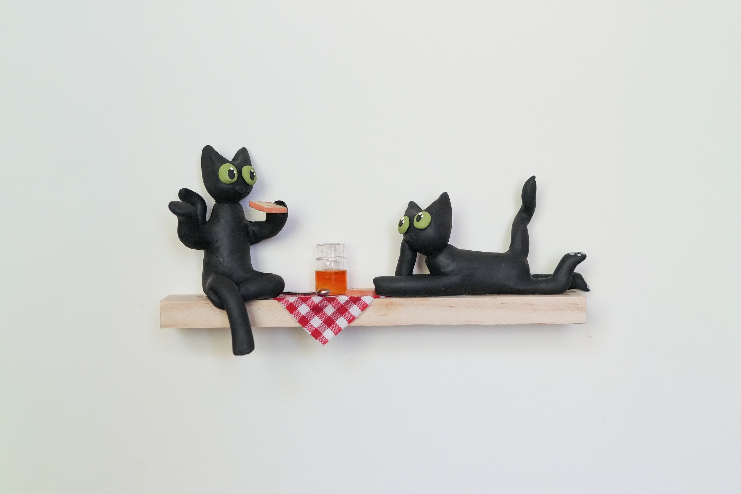 Two black cat figurines with large green eyes on a wooden shelf, one sitting with a pizza slice and the other laying down near a small jar of orange liquid, on a red and white checkered cloth against a plain cream wall.