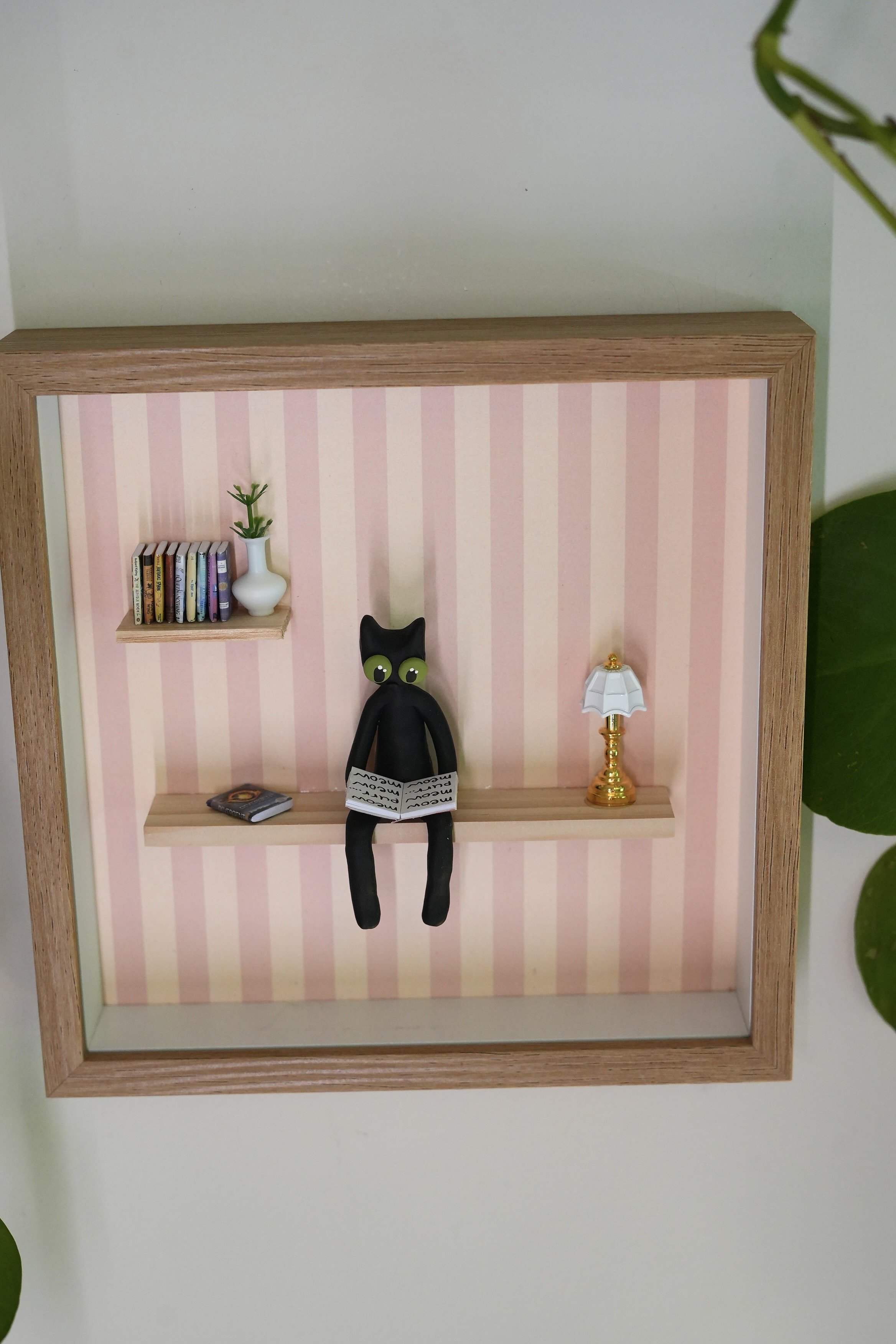 Miniature wall shelf with small books, a white vase with a green plant, a black cat figure holding a books, and a gold and white lamp, all against pink and white striped wallpaper.