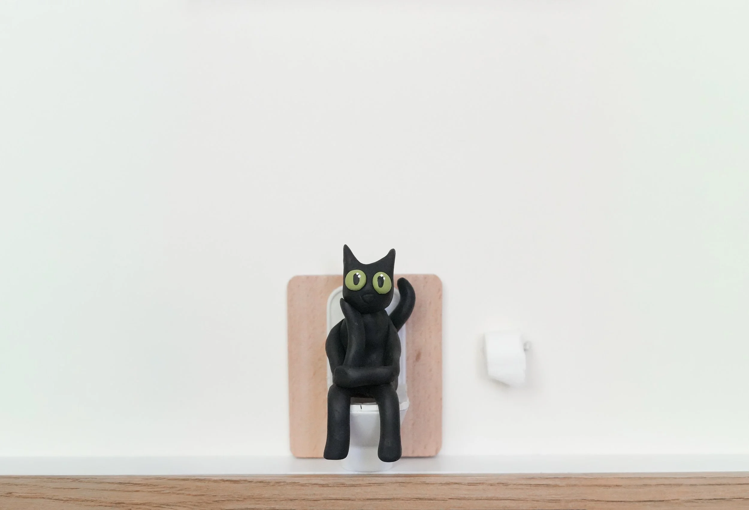 A black clay figure with large green eyes, sitting on a toilet and waving with one hand, against a white wall with a light pink bathroom tile behind it.
