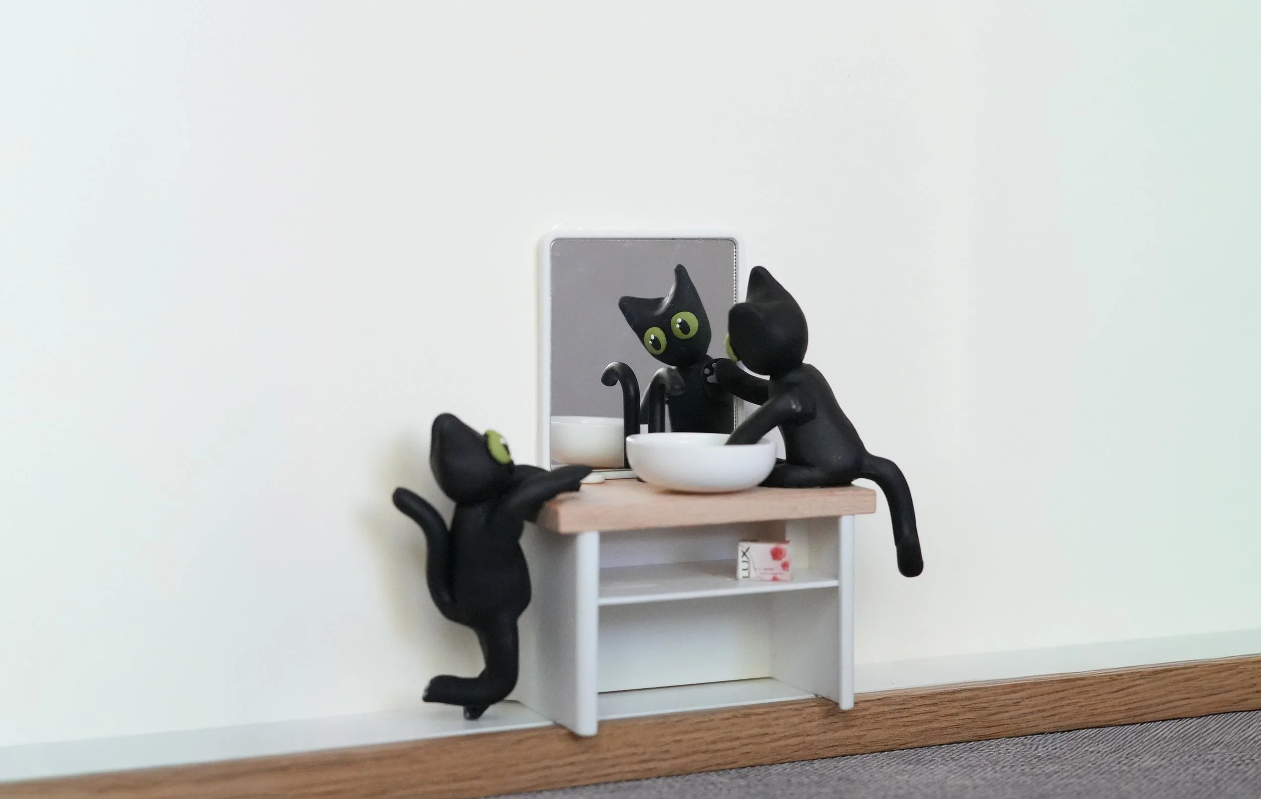 Miniature black cat figurines with green eyes sit at a small bathroom vanity with a mirror, sink, and soap dispenser.