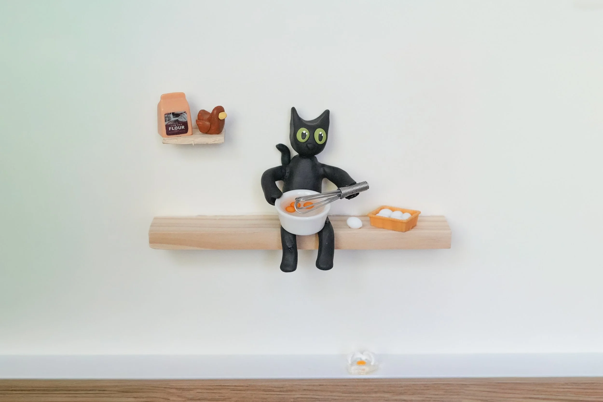Miniature black cat holding a whisk and shopping for eggs while sitting on a wooden shelf, with a bowl of eggs and a basket of eggs nearby, and a small shelf with flour and a chicken on the wall.