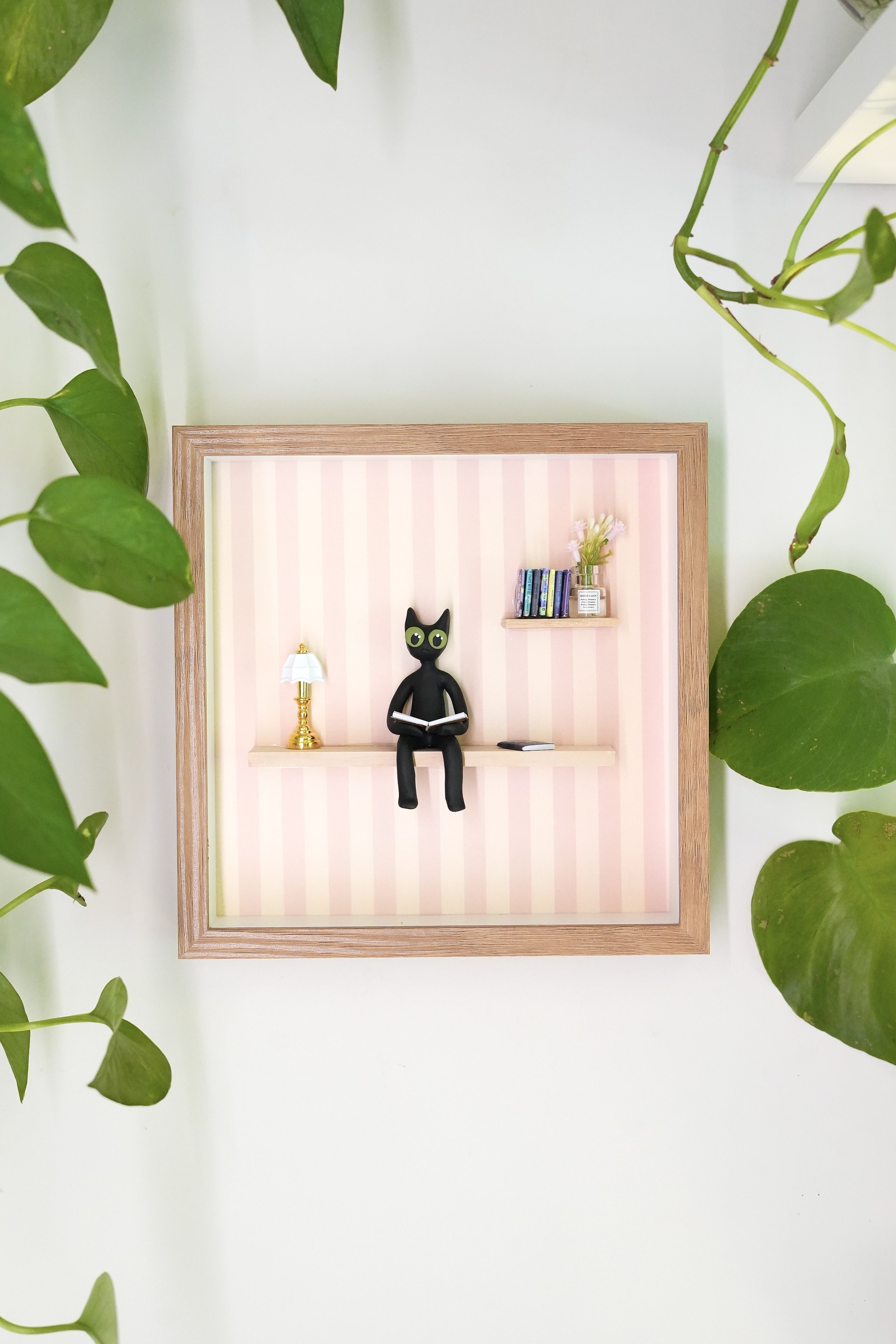 Decorative wall shelf with books, flowers, a black cat figurine reading a book, and a small lamp, framed by green leafy plants.