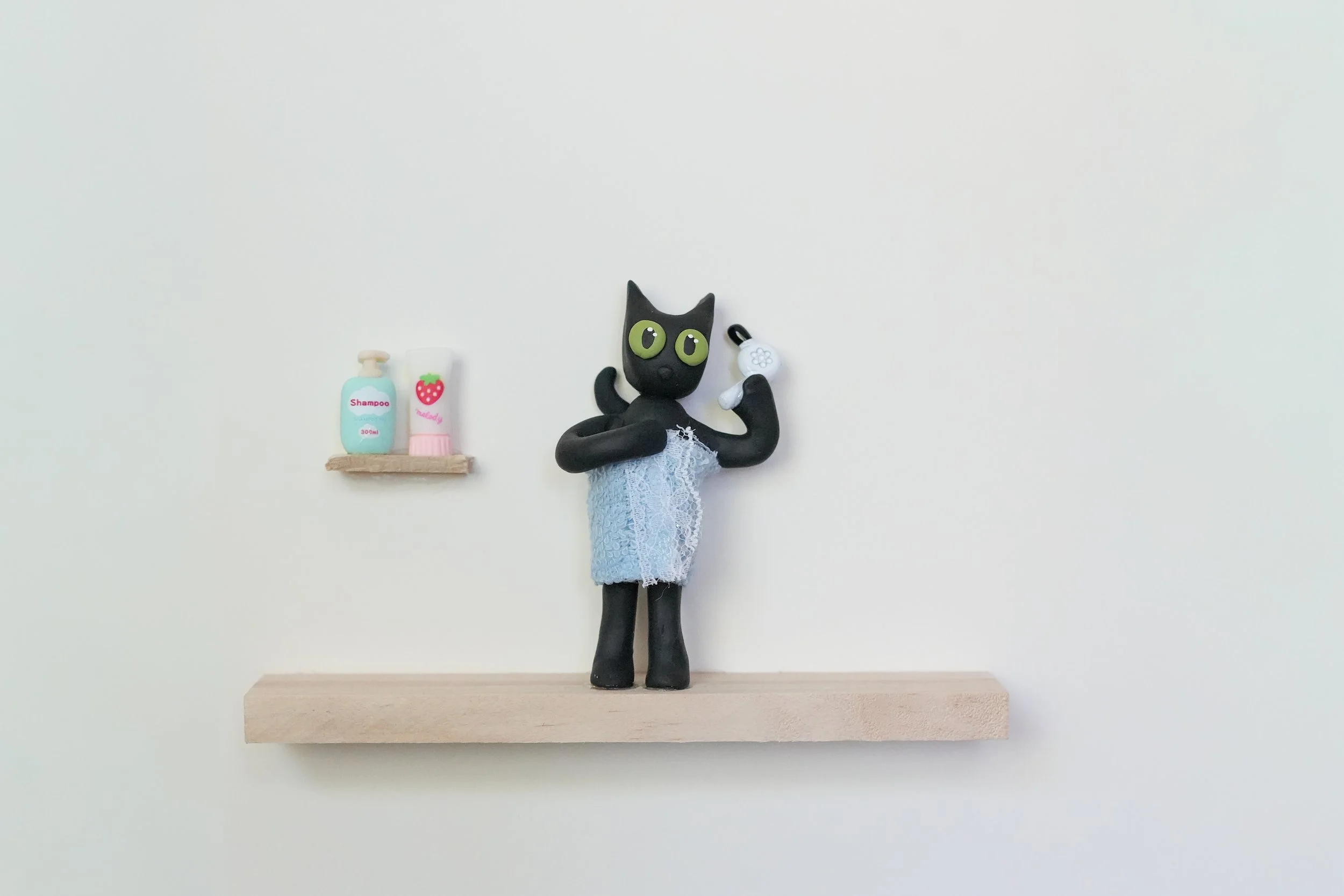 A black cat figurine with large green eyes, wearing a blue lace dress, holding a white telephone receiver to its ear, standing on a light wooden shelf against a plain white wall. To the left of the figurine, there is a small shelf with two bottles of