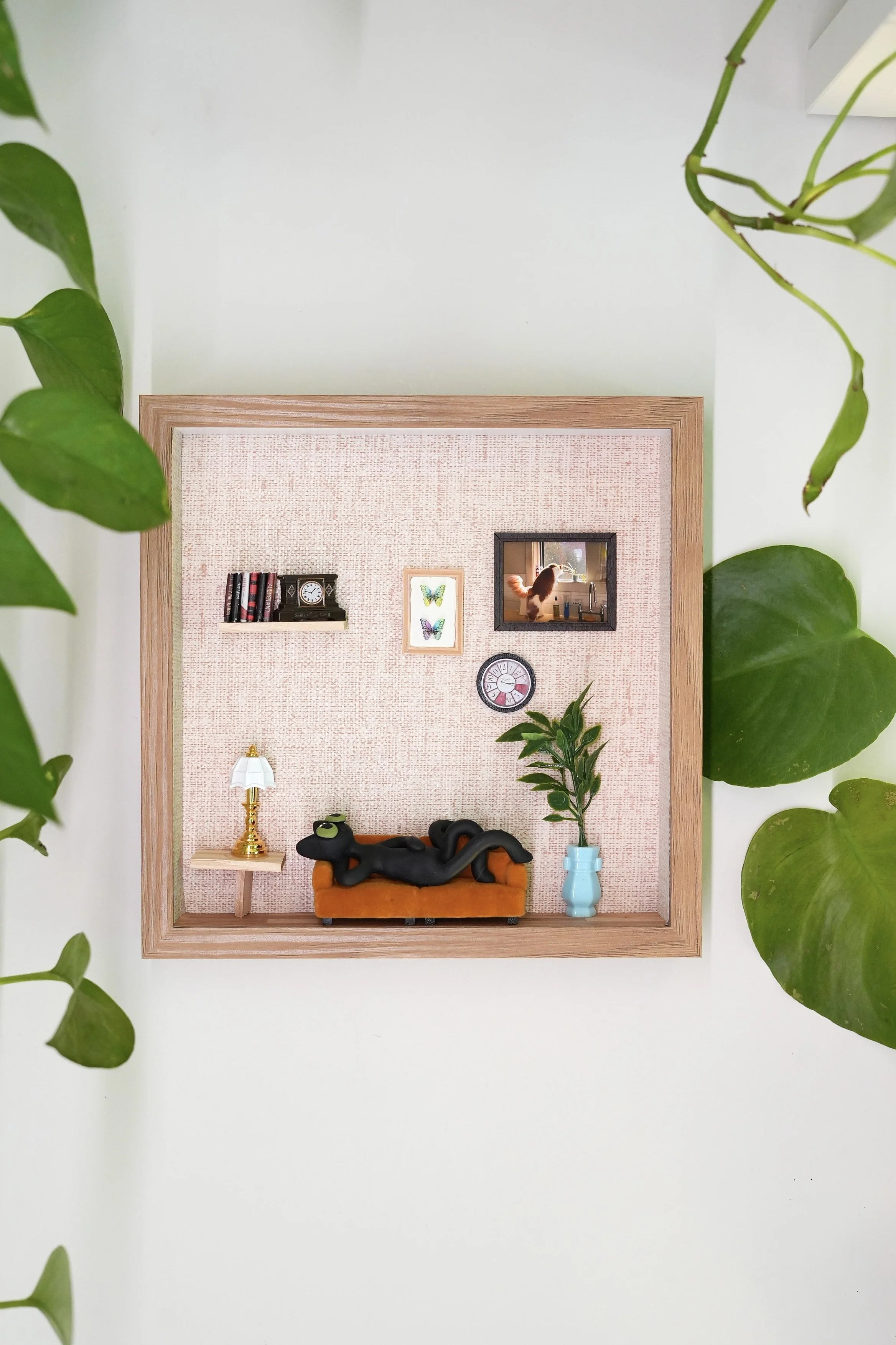 Miniature room with a pink textured wall, decorated with two framed butterfly art pieces, a small clock, a black cat statue lying on an orange couch, a green plant in a blue pot, a gold-lamp table, and wall shelves with books, a clock, and a small ca