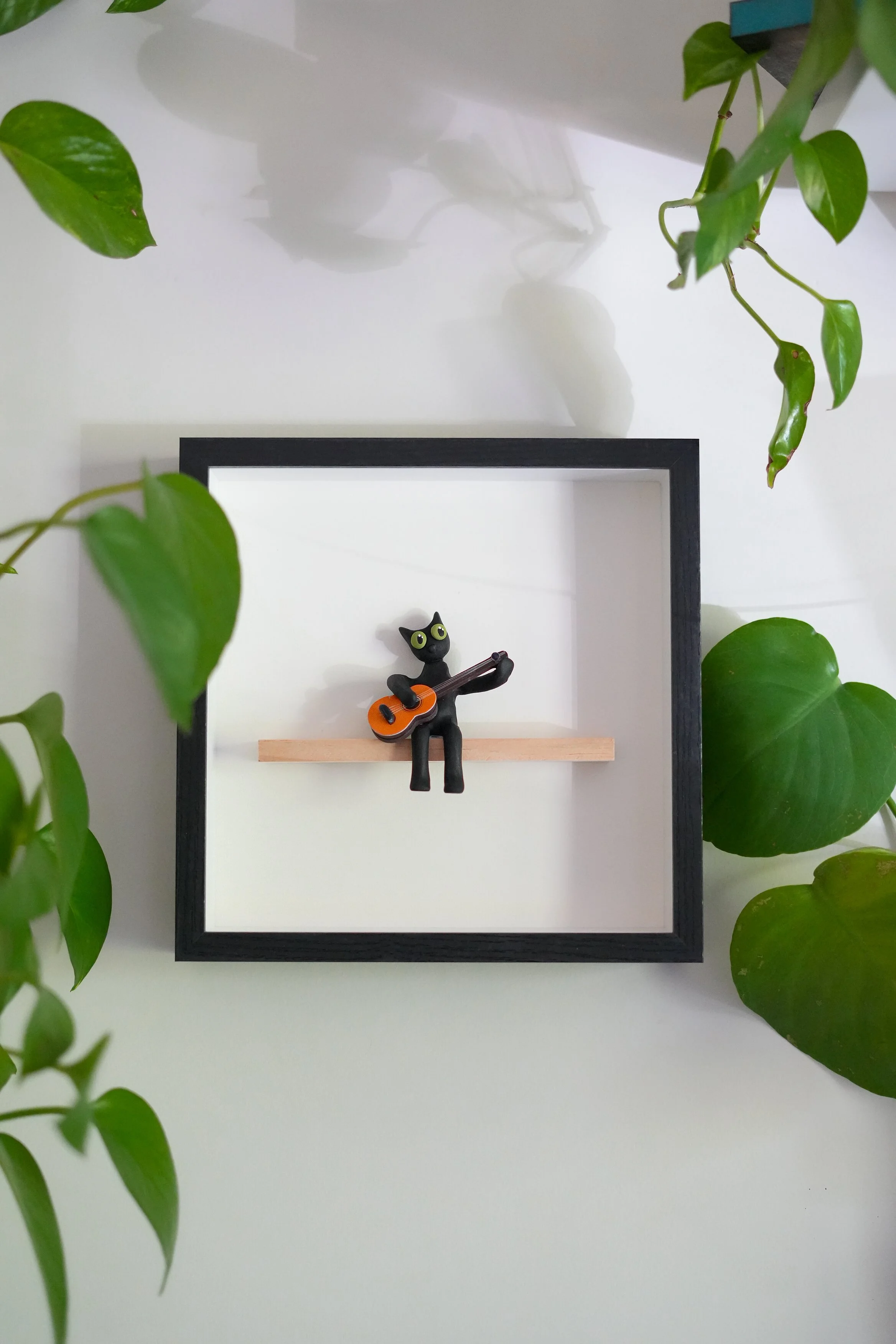 A small black cat figurine with green eyes playing an orange guitar, placed inside a black square frame with a white background, surrounded by green houseplants.