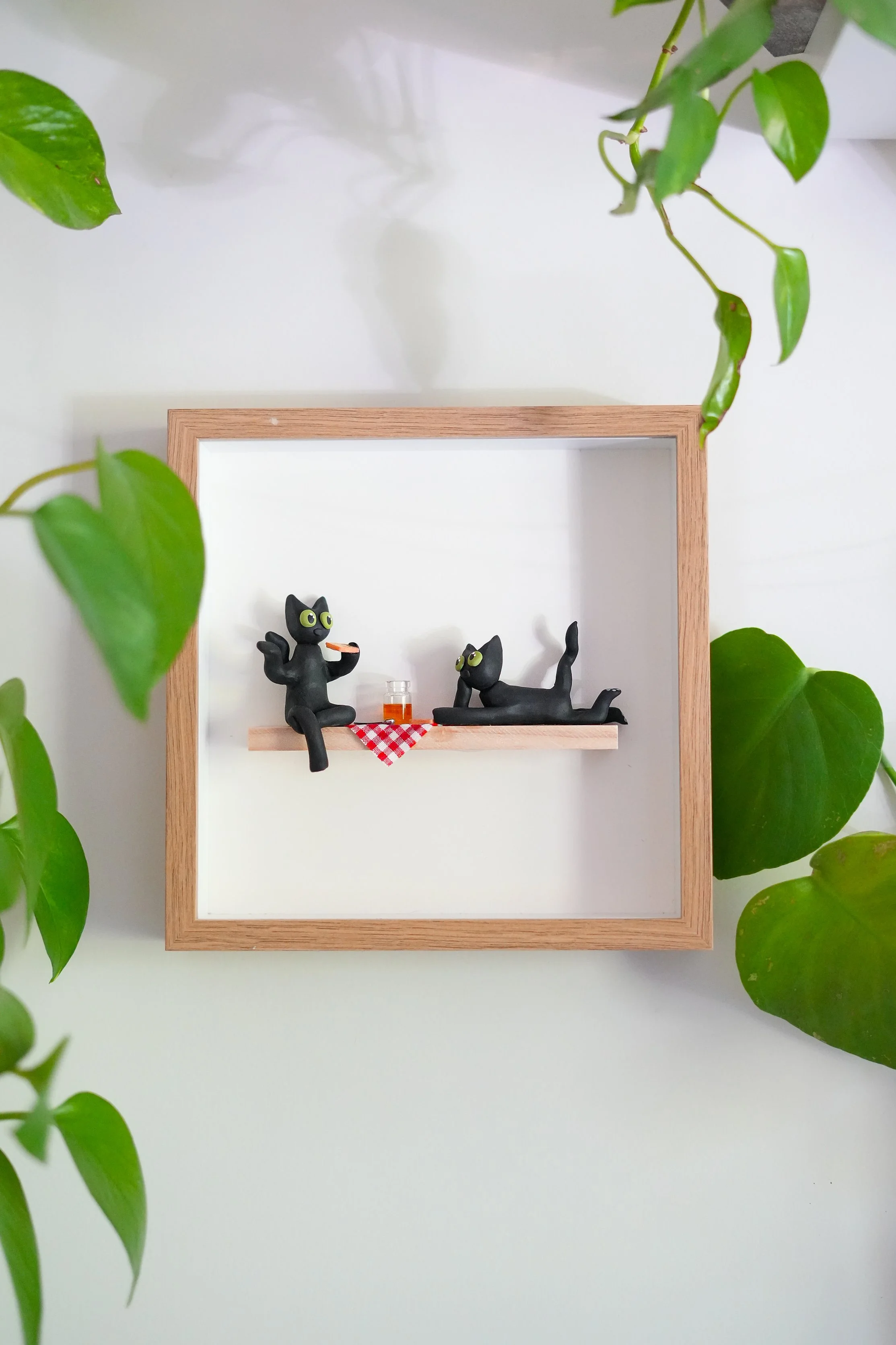 Decorative display of two black cat figurines inside a wooden shadow box, with one sitting and the other lying down, a small glass of orange beverage, and a red checkered napkin on a shelf, surrounded by green leaves.