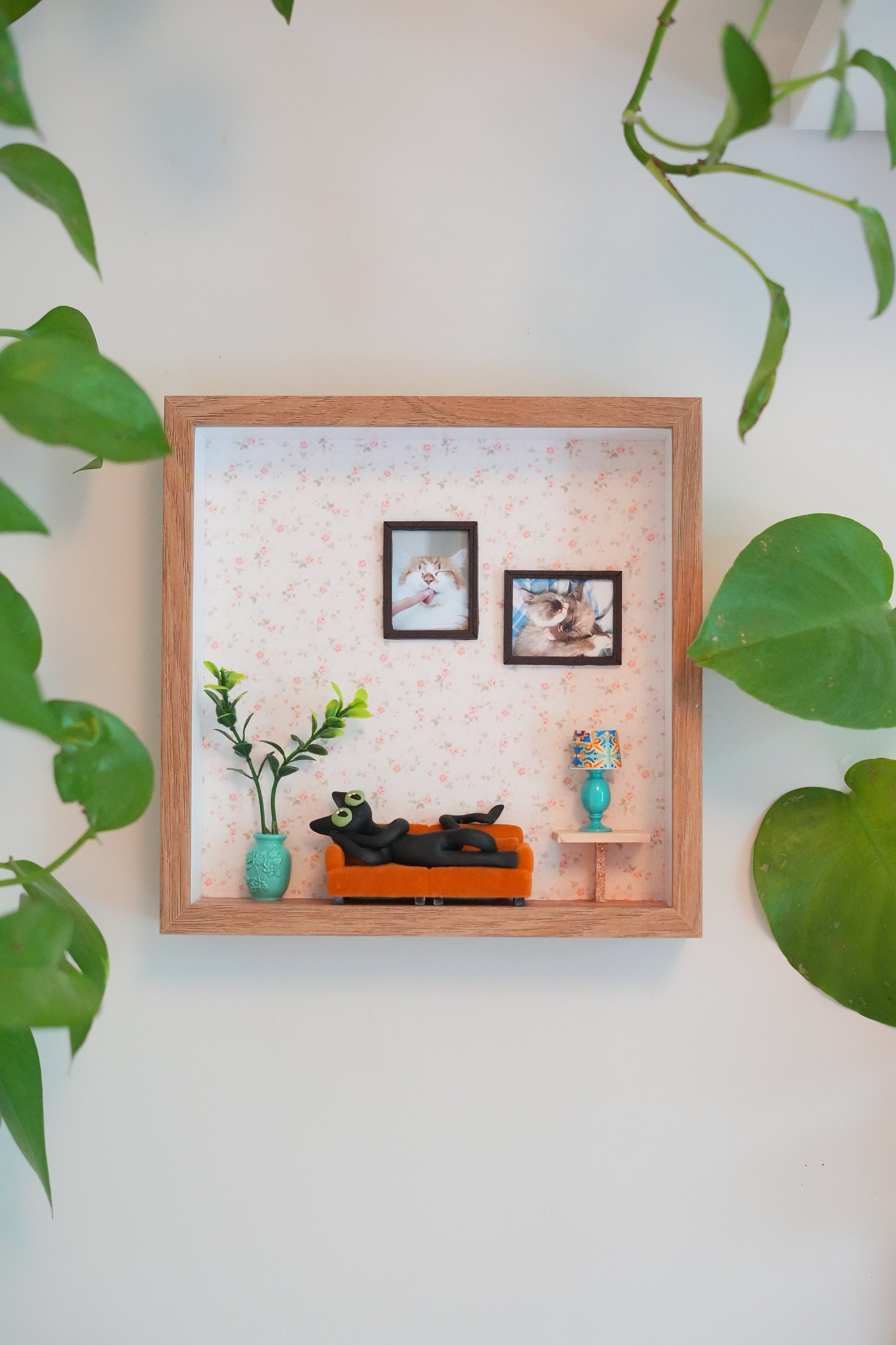 Miniature framed scene with two small photographs of cats, arranged in a decorative setting with plants, a teal vase, and a small table with a colorful lamp.