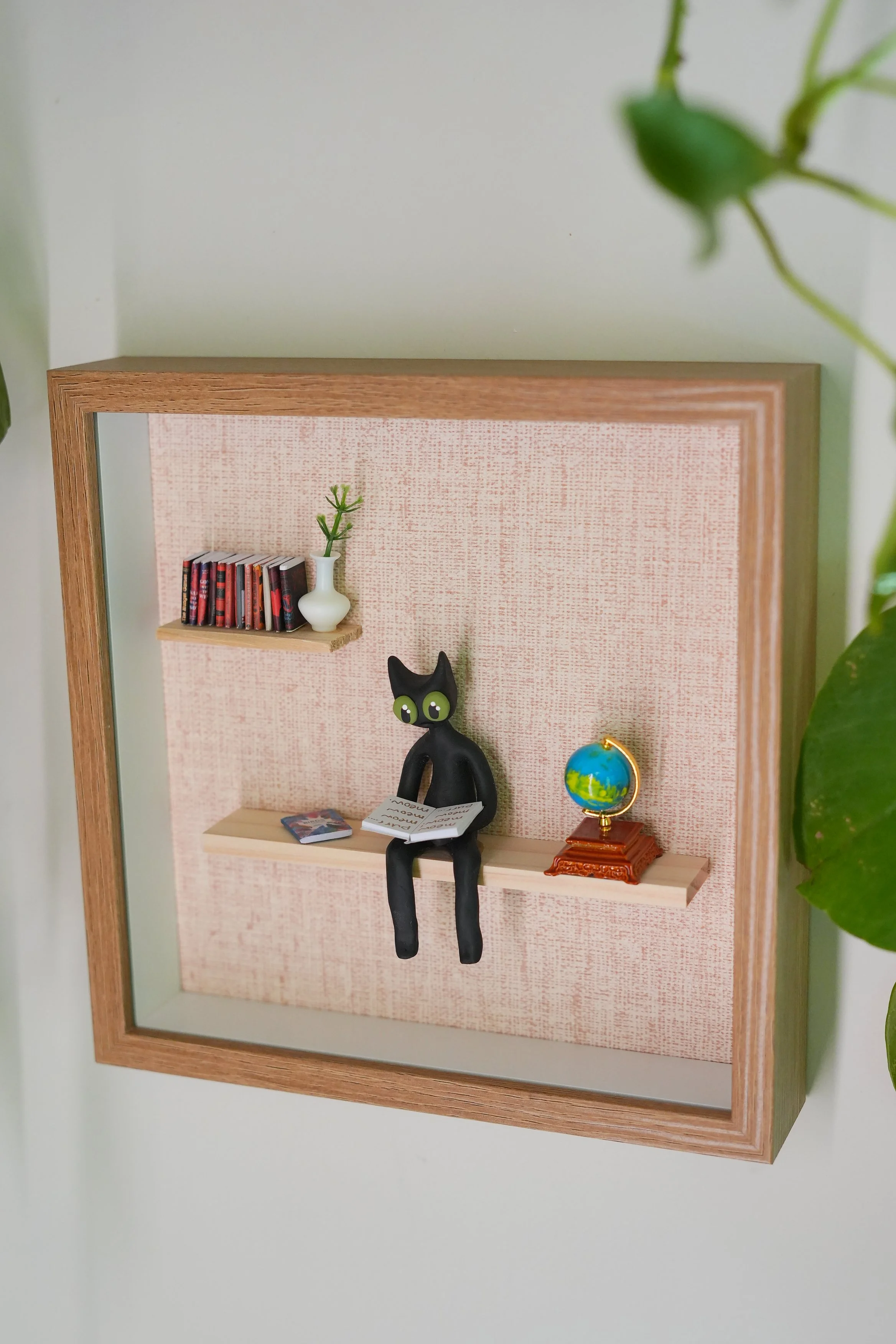 Miniature black cat figurine with green eyes sitting on a small shelf, with a globe and some books nearby, against a pink textured background inside a wooden frame