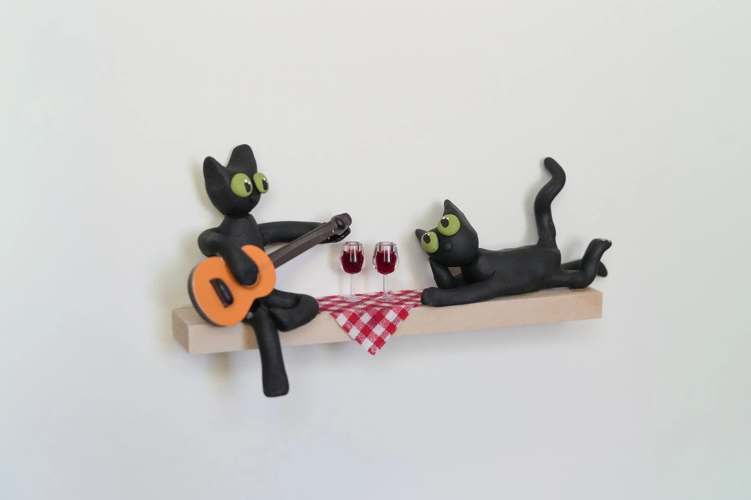 Two black cat figurines on a floating shelf, one sitting with an orange guitar, the other lying down with two glasses of red wine and a red checkered cloth underneath.