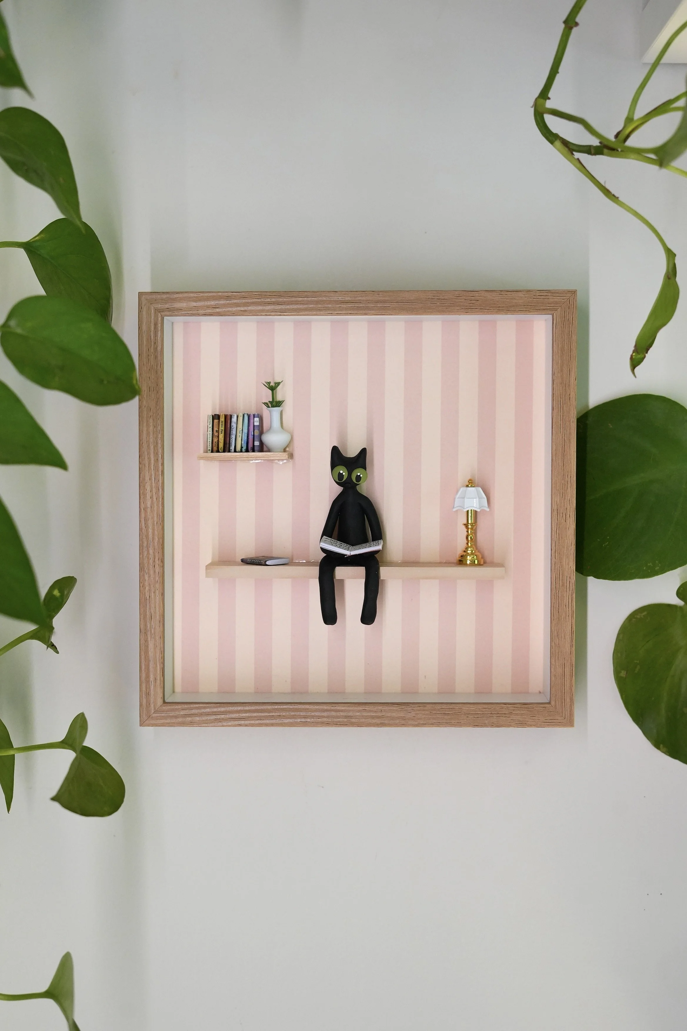 Miniature framed diorama with a black cat figure reading a book, surrounded by a small bookshelf, a white vase with a plant, and a lamp, against pink striped wallpaper, framed by green leaves.
