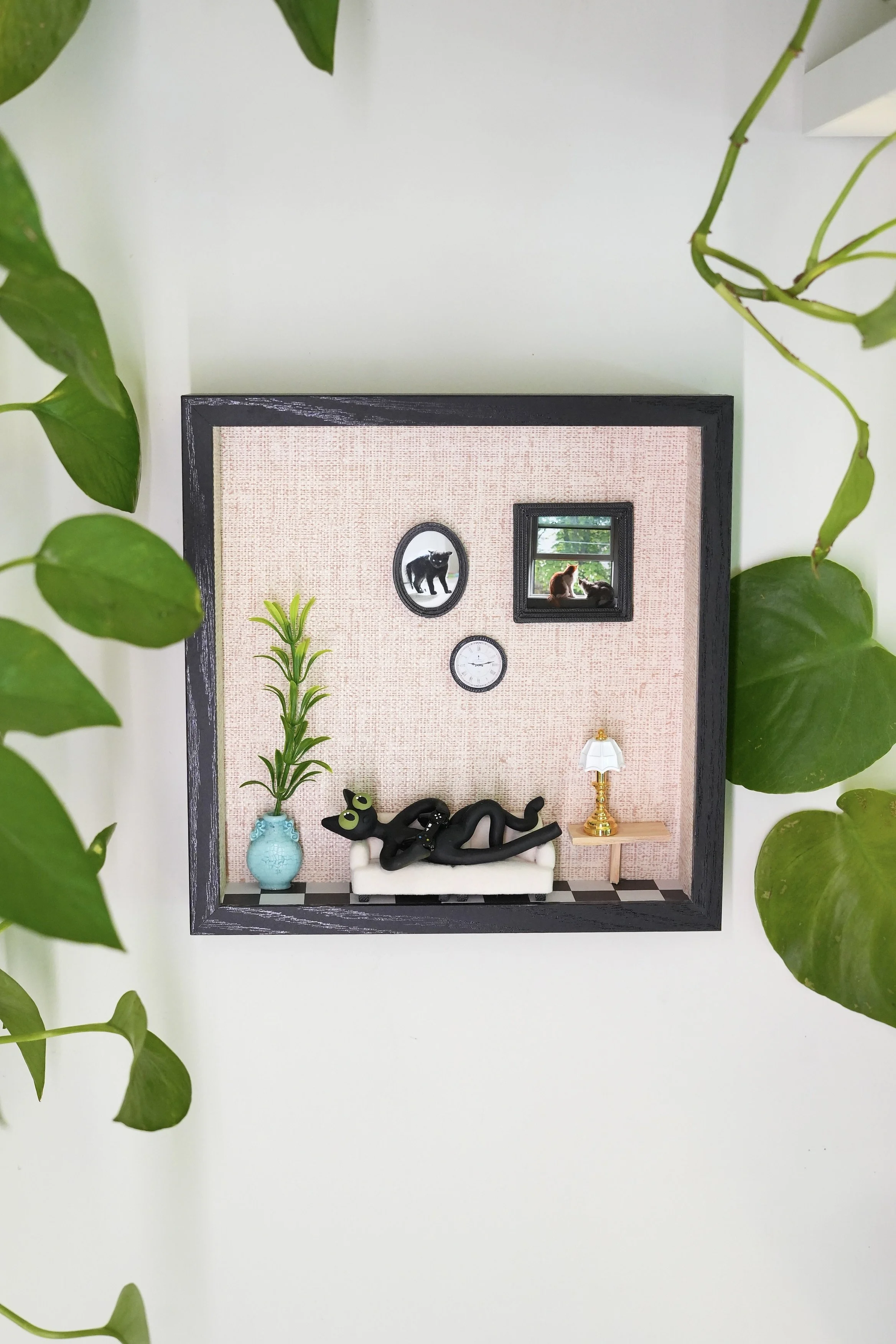 Decorative framed miniature room scene with a sofa, black cat figurines, a small table with a lamp, a potted plant, and wall accents including a small mirror and clock, surrounded by green leafy plants.