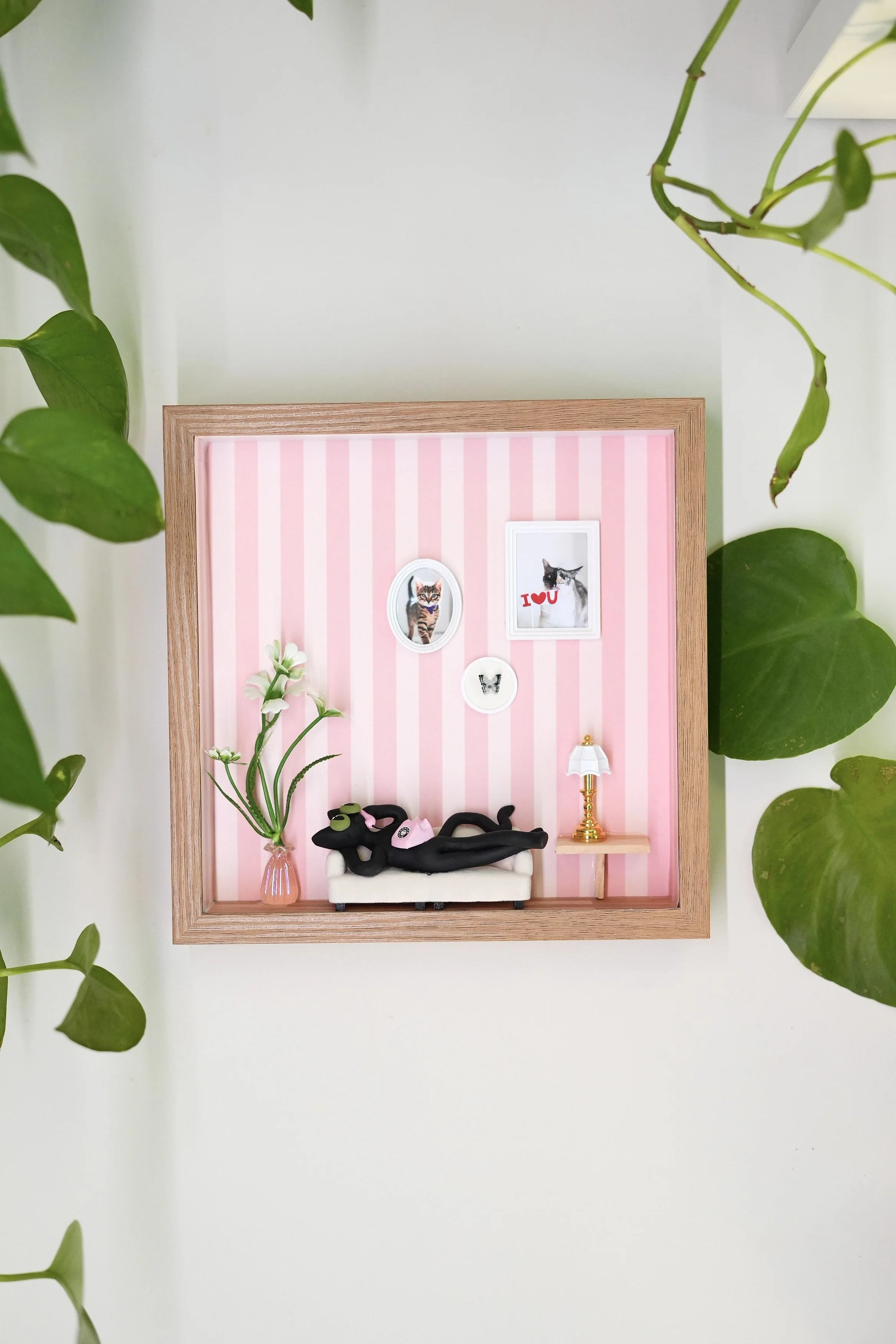 Decorative shadow box featuring miniature cat figurines, framed pictures of cats, a small lamp, and flower decorations, surrounded by green house plants.