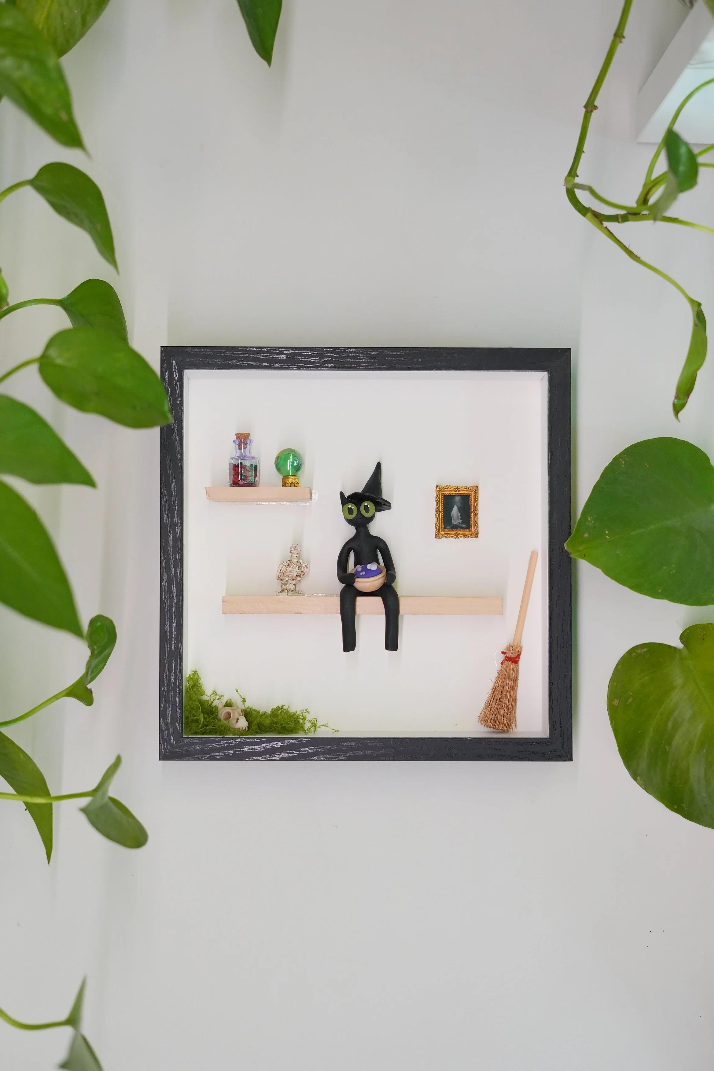 Decorative wall display with small shelves in a black frame, featuring a witch figurine, a small framed picture, a skeleton ornament, a green glass ball, a jar with red items, moss, and a miniature broom, surrounded by green leafy plants.