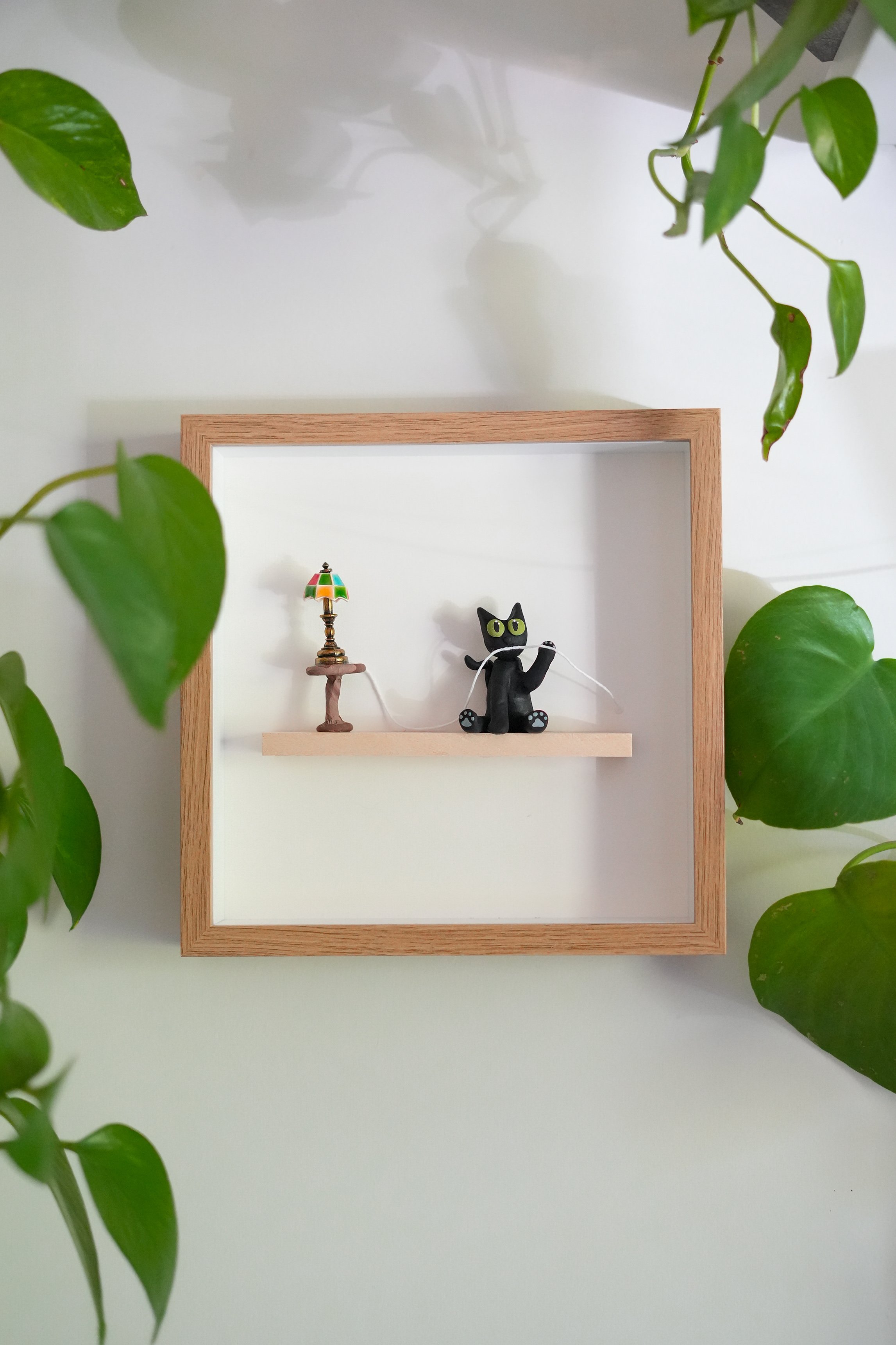 Decorative shadow box with a small colorful lamp and a black cat figurine holding a white string, surrounded by green houseplants.