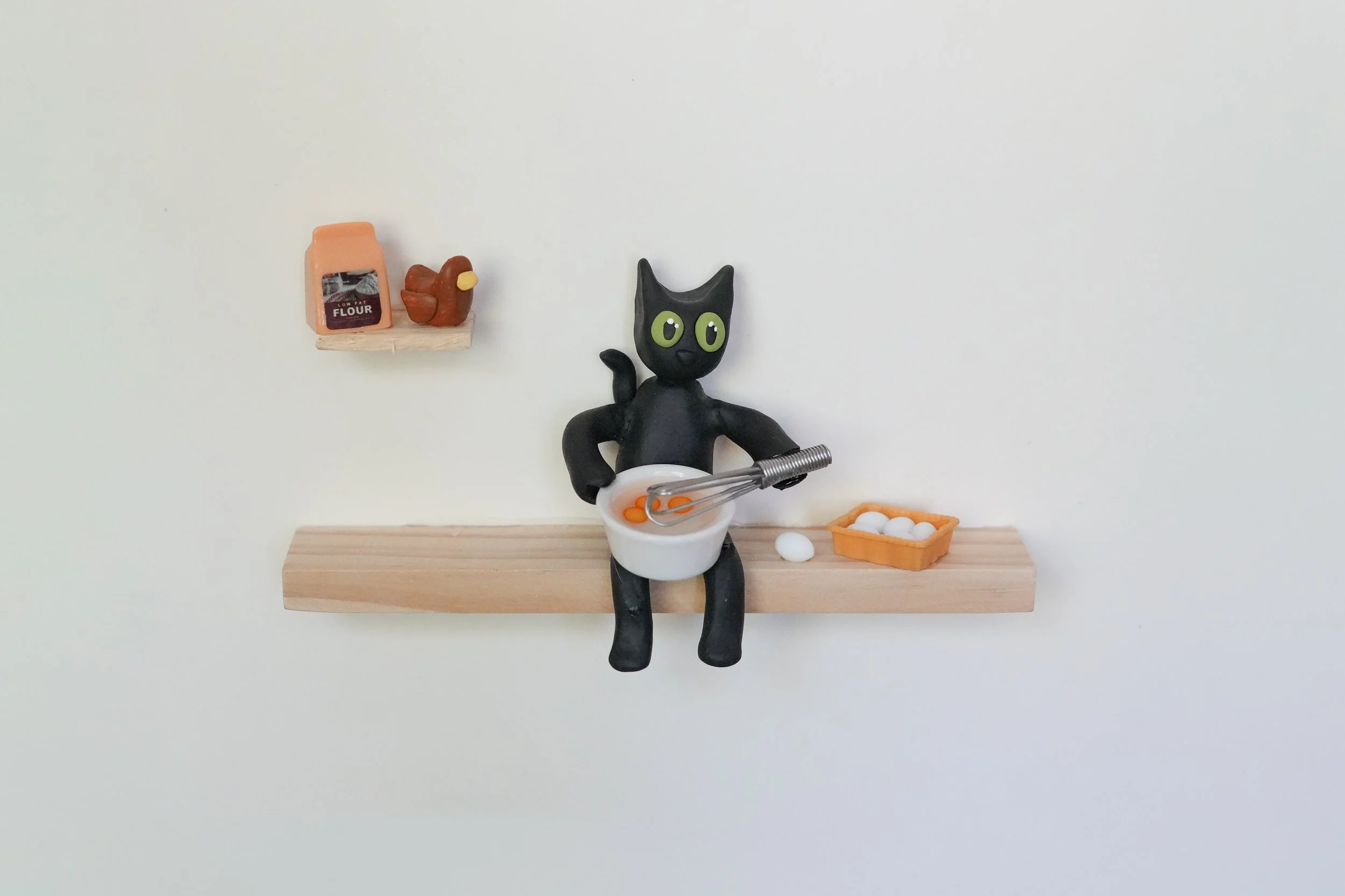 A small figurine of a black cat with green eyes holding a mixing bowl and whisk, sitting on a wooden shelf with a basket of eggs and a tiny shelf with a bag of flour and a chicken model.
