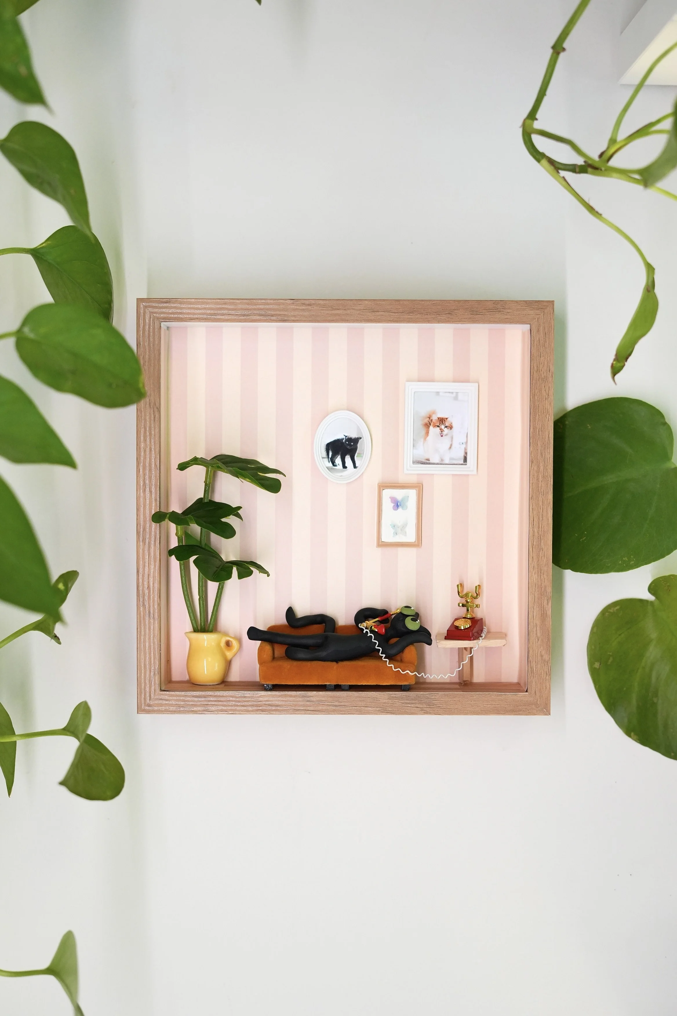A shadow box with a pink striped background containing small framed pictures of a black cat, a fox, and butterflies on the wall, a yellow vase with green plant, a miniature orange sofa with a black cat sculpture, and other small decorative items, sur