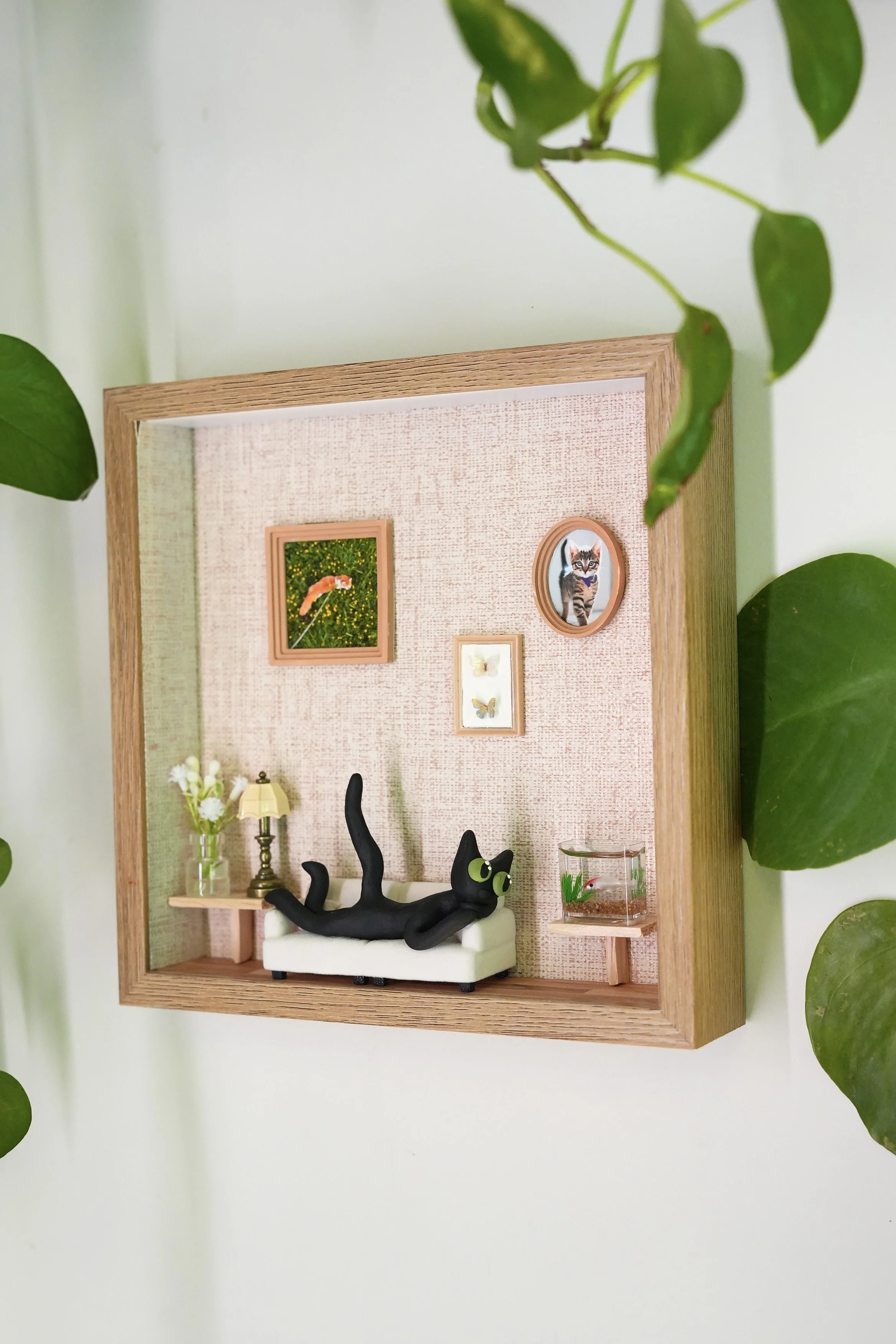 Miniature room scene with a black cartoon cat figurine lounging on a tiny white sofa, bookshelf with a lamp, flower vase, and a glass fish tank, decorated with framed pictures of a cat, a woman, and butterflies, surrounded by green leafy plants.