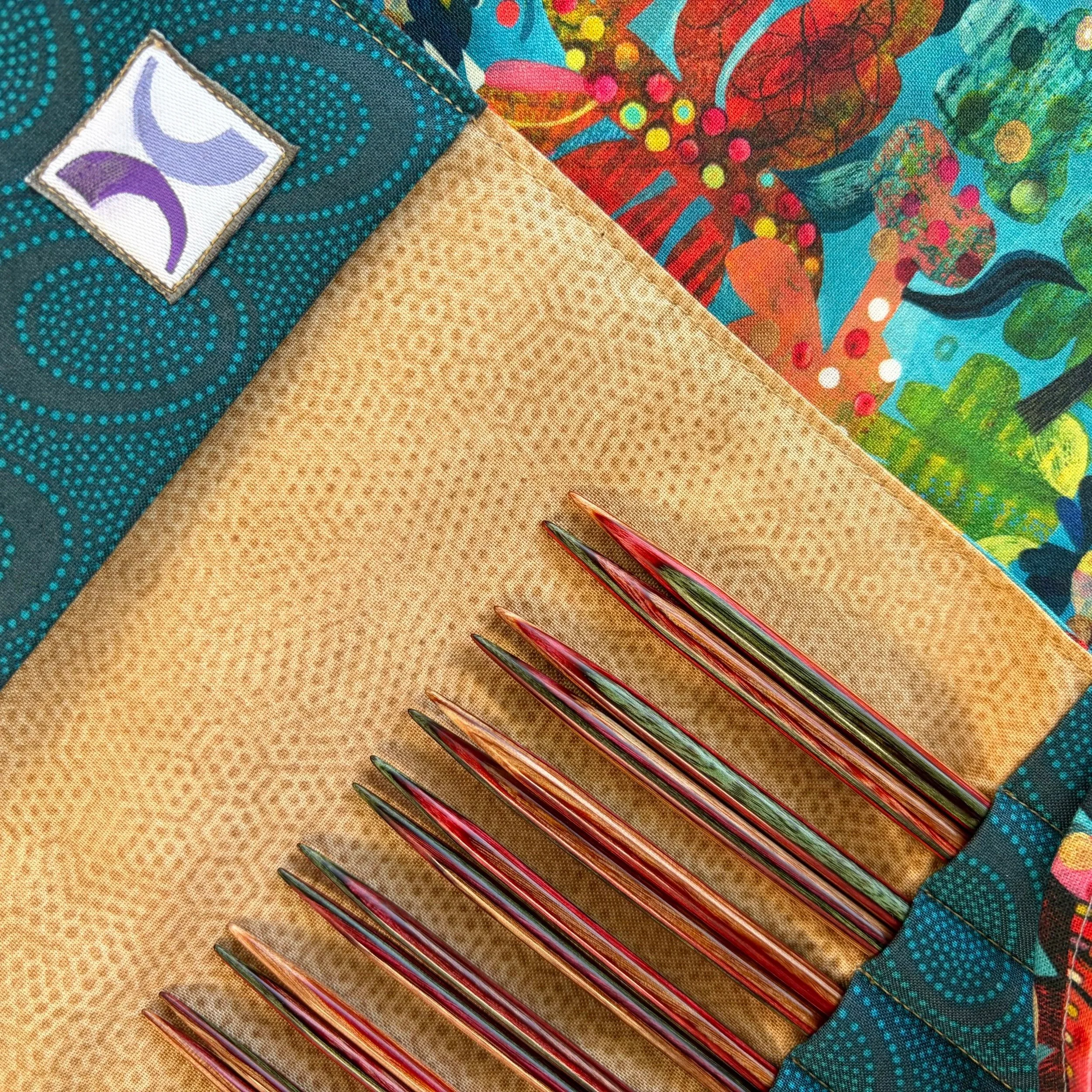 Sagebrush Darn Pretty™ knitting needle set. Needles are shades of natural, orange, and sage green in a case with teal pockets, gold background, and outside fabrics of colorful plants and crows.