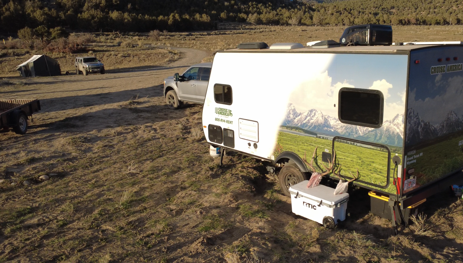 Camping setup in a desert landscape with a white travel trailer, nearby trucks, and a tent in the background. The trailer has a mountain and field mural, and there is hunting antlers and RTIC cooler in front.